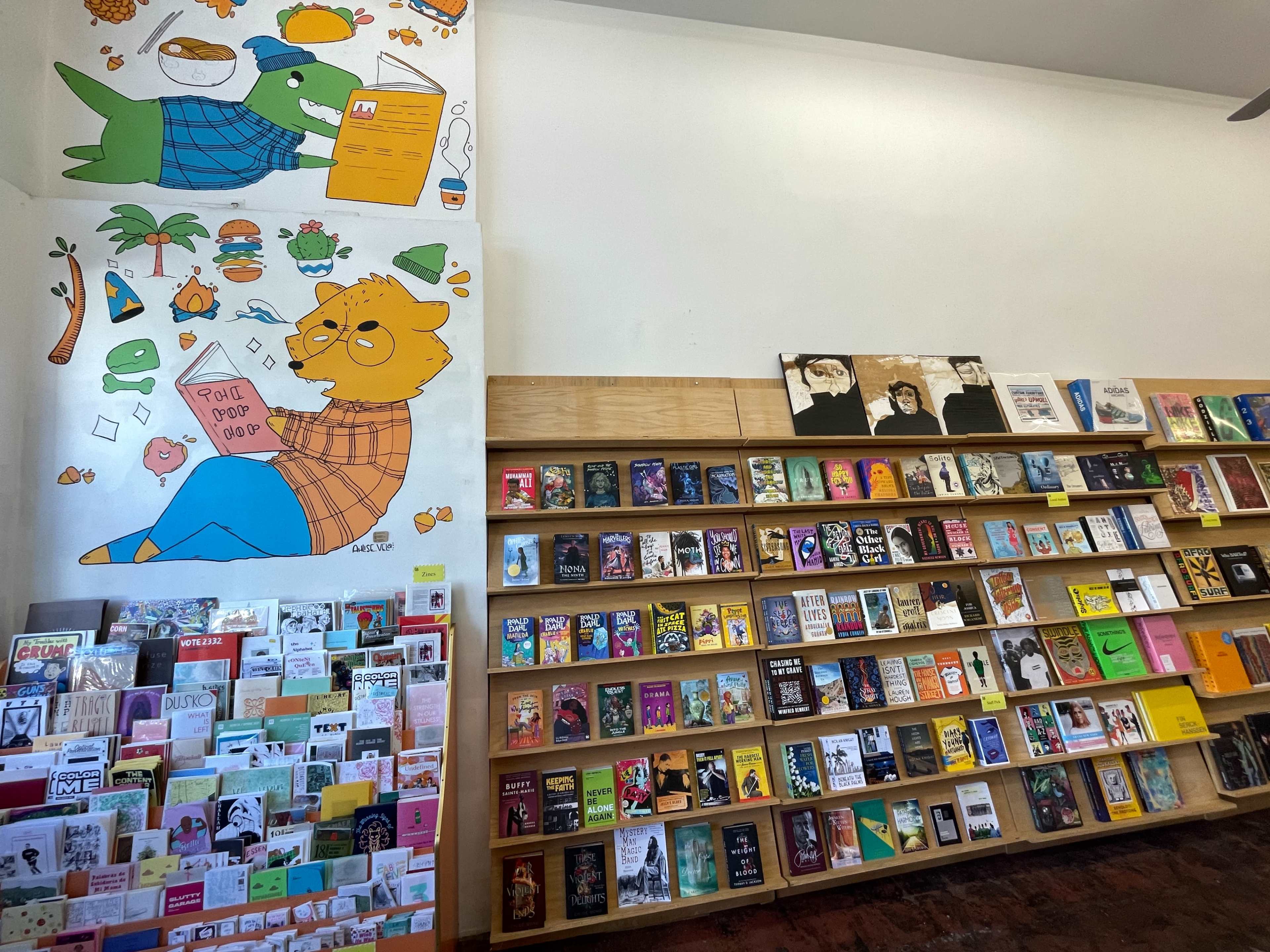 The image shows a cozy bookstore with a mural of cartoon animals on the wall and shelves filled with various books and greeting cards.