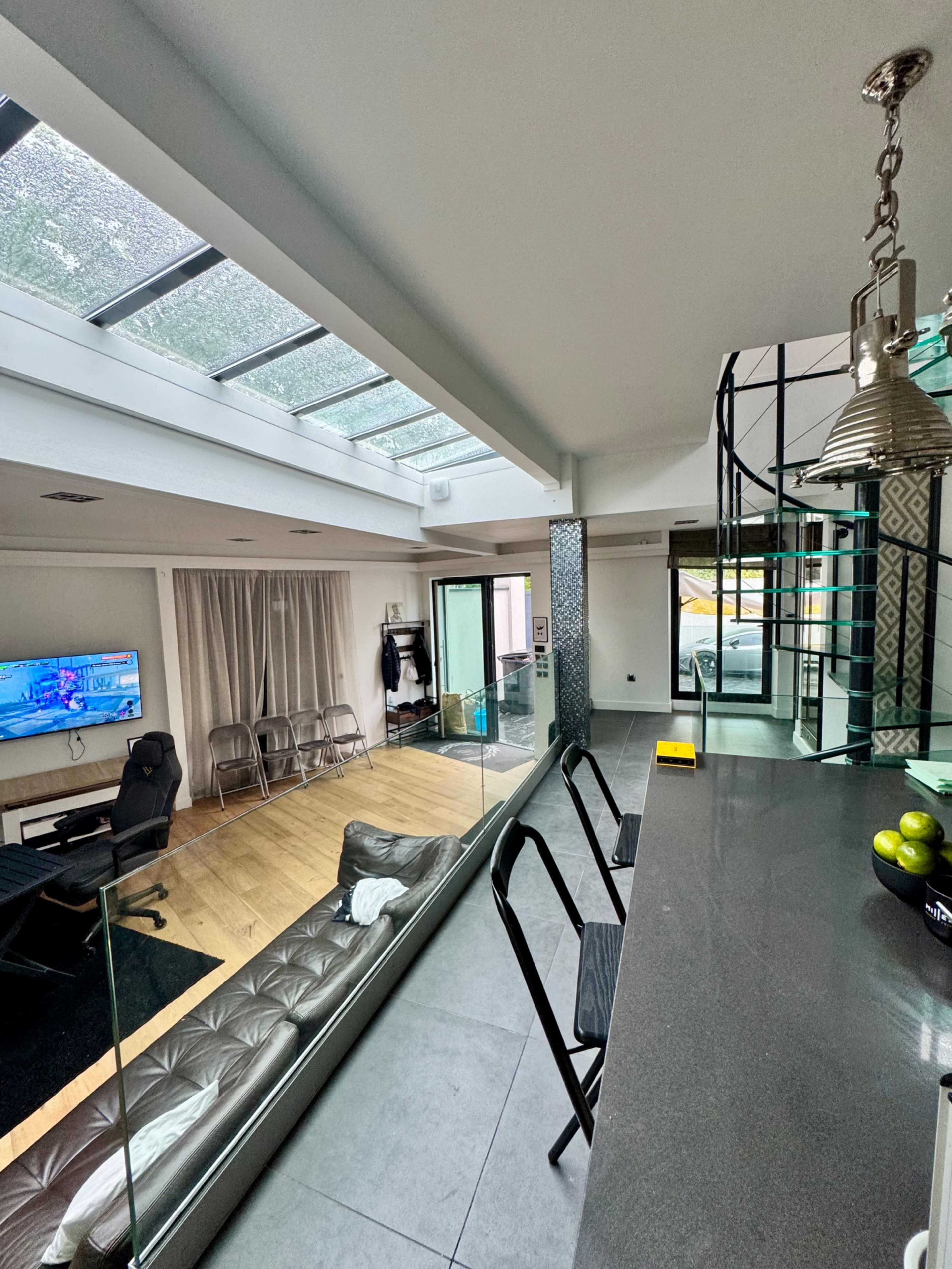 The image shows a modern open-concept living space with a glass ceiling, a kitchen counter, a living area with a TV, and a spiral staircase leading to another level.