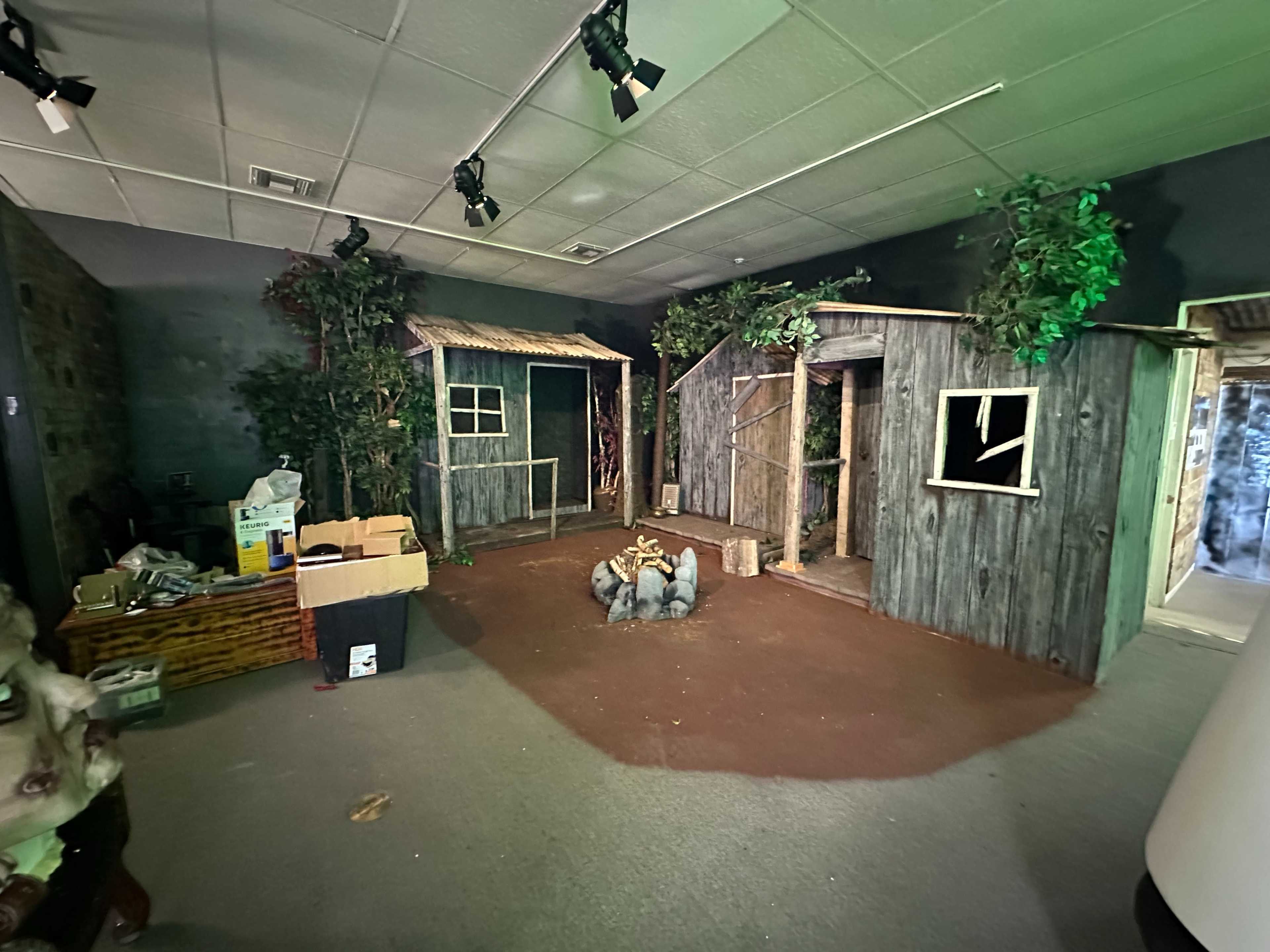 The image shows a dimly lit room designed to resemble a rustic outdoor setting with two wooden structures, surrounded by artificial vegetation and a central fire pit.