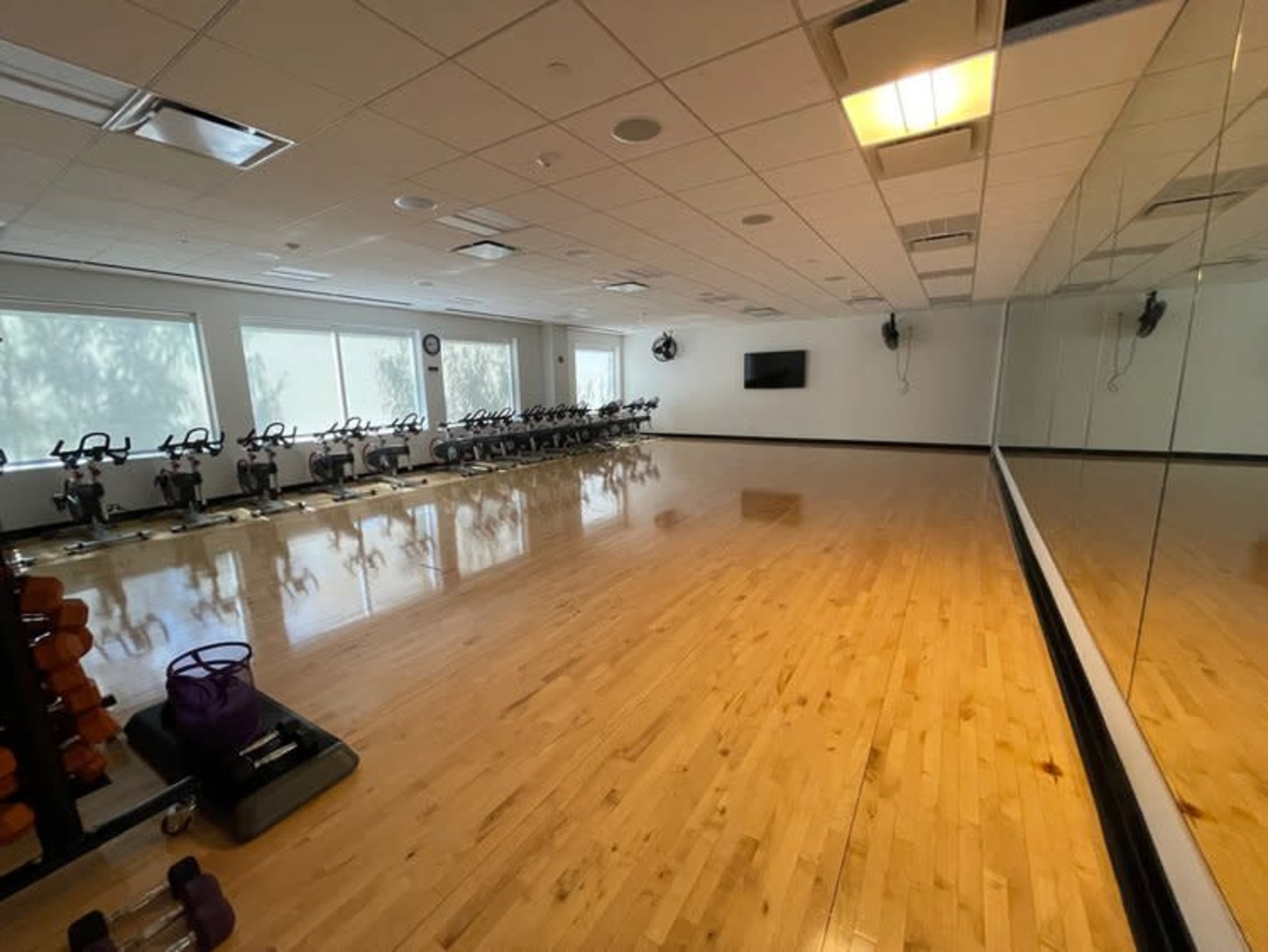 The image shows a spacious, well-lit fitness studio with wooden flooring, exercise bikes lined up against one wall, and a large mirror along another wall.