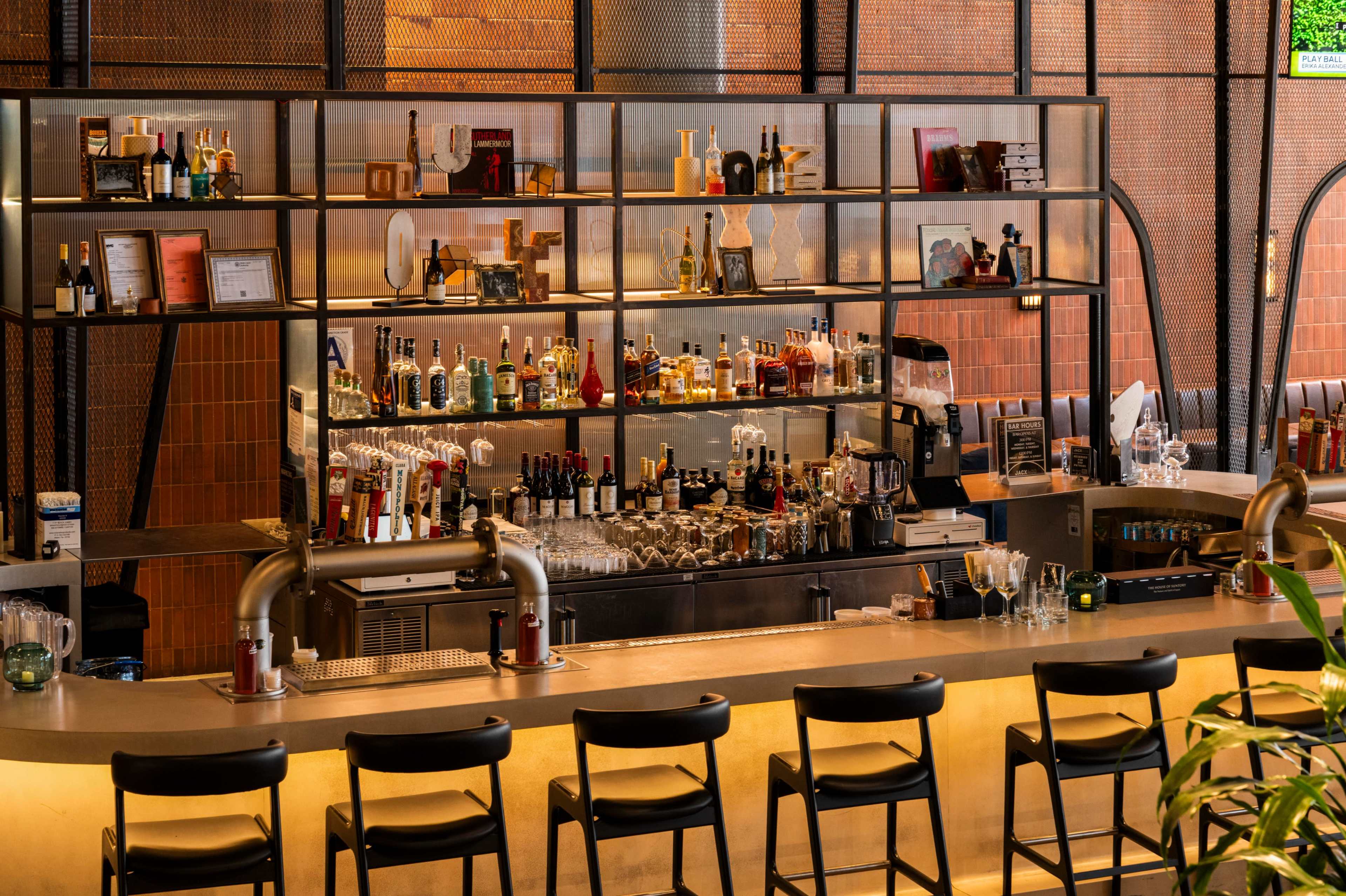 The image shows a modern bar with a well-stocked shelf of various liquor bottles and a long countertop lined with black chairs.