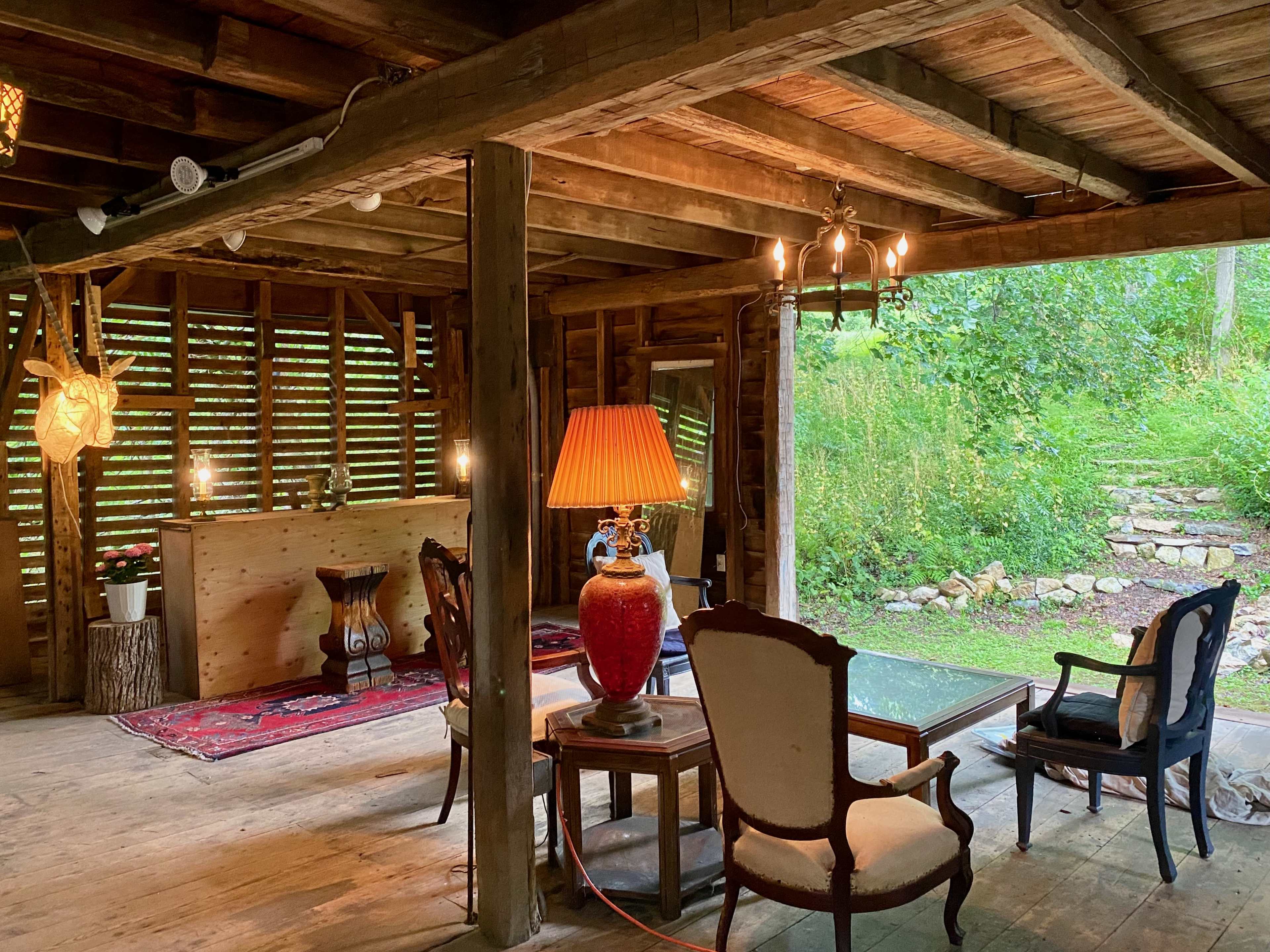 The interior of a rustic wooden cabin features a seating area with chairs and a table, illuminated by a lamp, and a view of a garden through large windows.