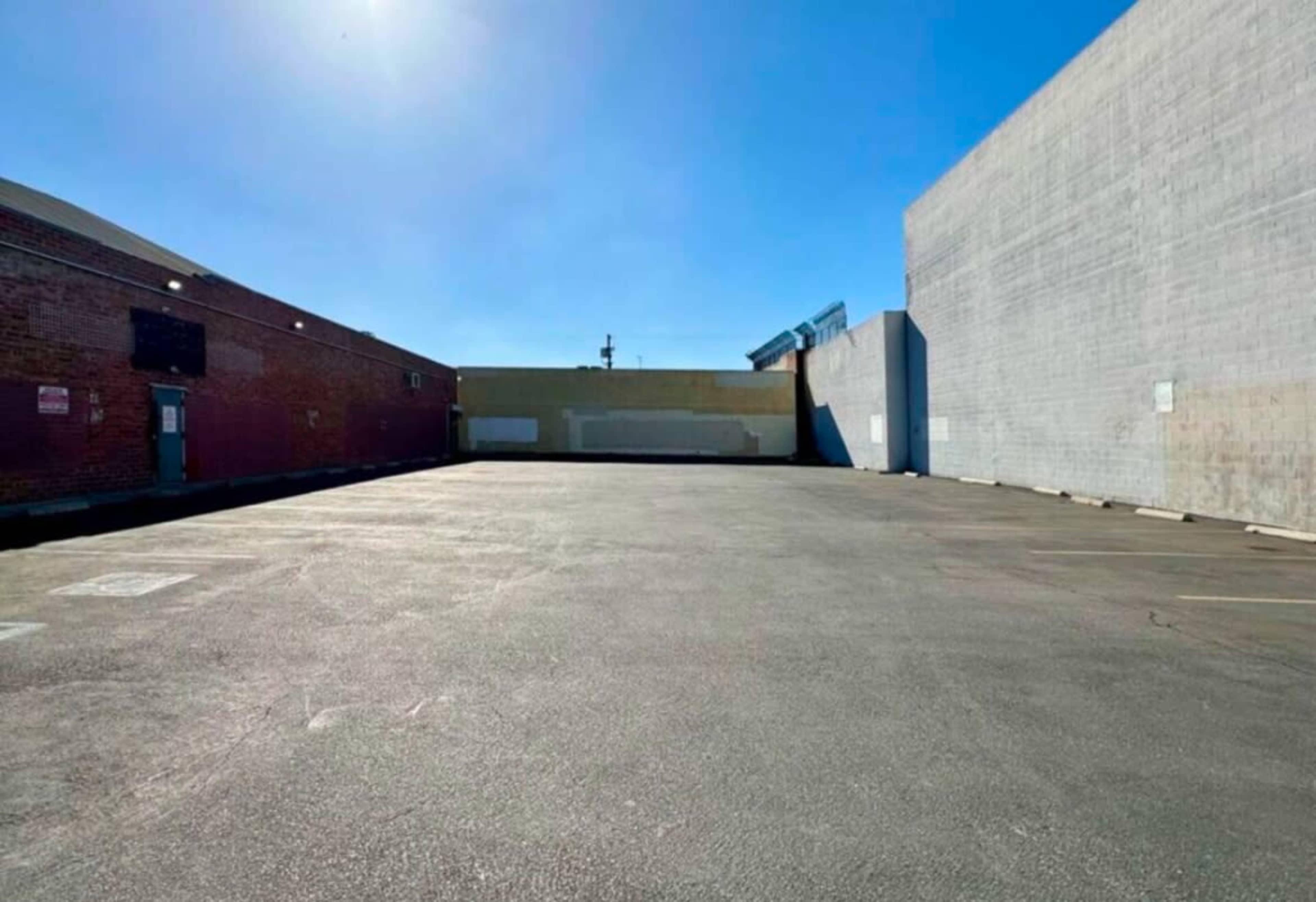The image shows a vacant asphalt parking lot surrounded by brick and concrete buildings under a clear blue sky.