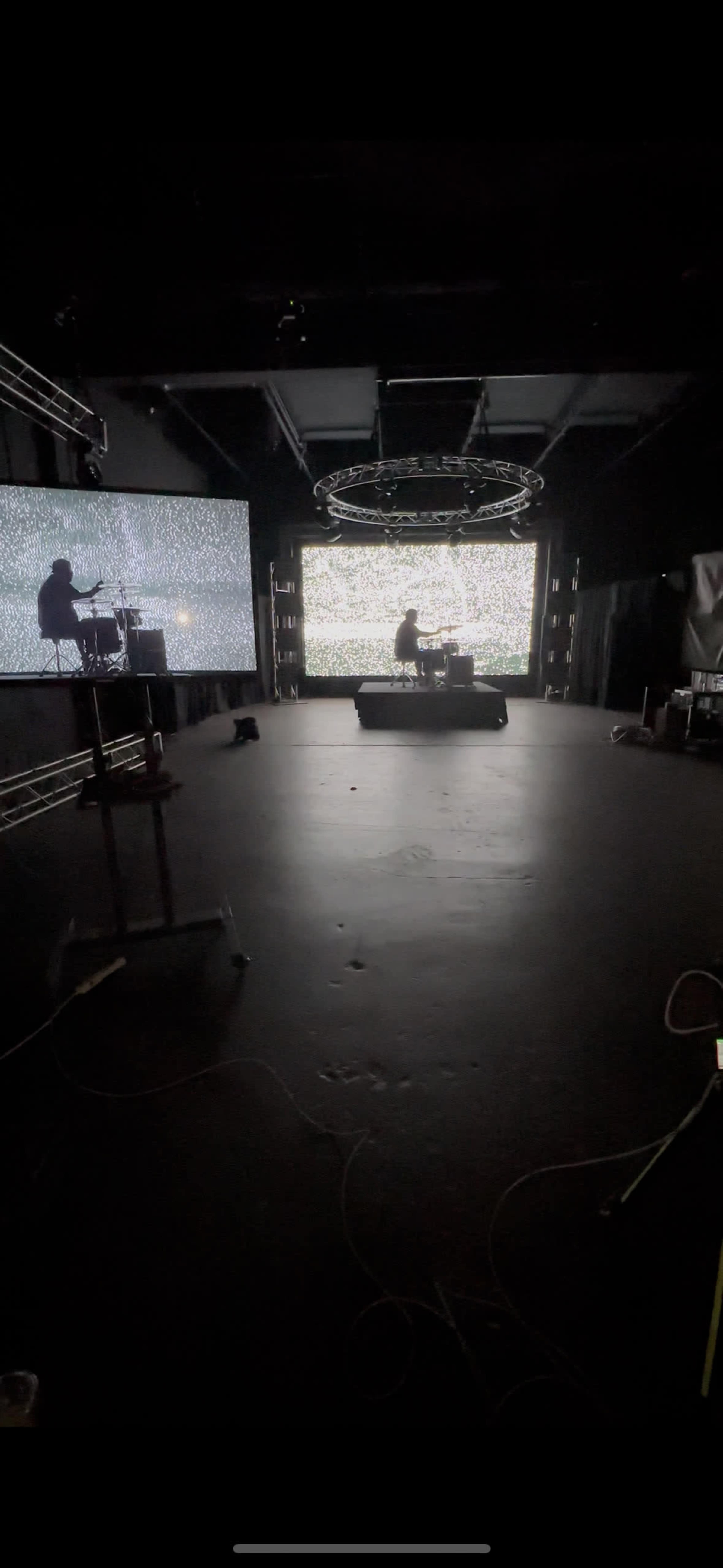 The image shows a dimly lit performance space with two large screens displaying static, and a person performing on a stage in the background.
