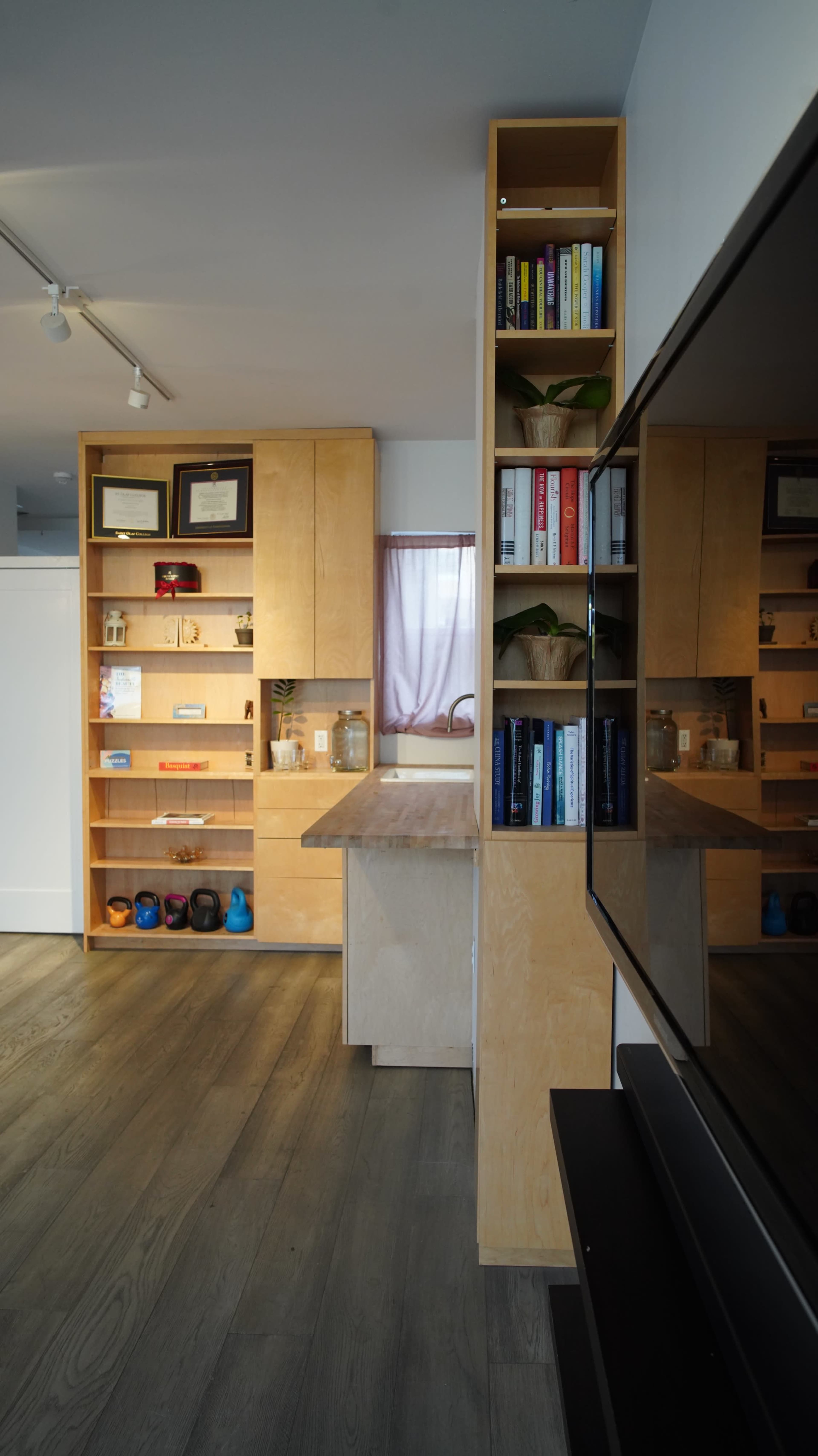 The image shows a modern office space with wooden shelving units displaying various items, a television on the right, and a small desk in the center.