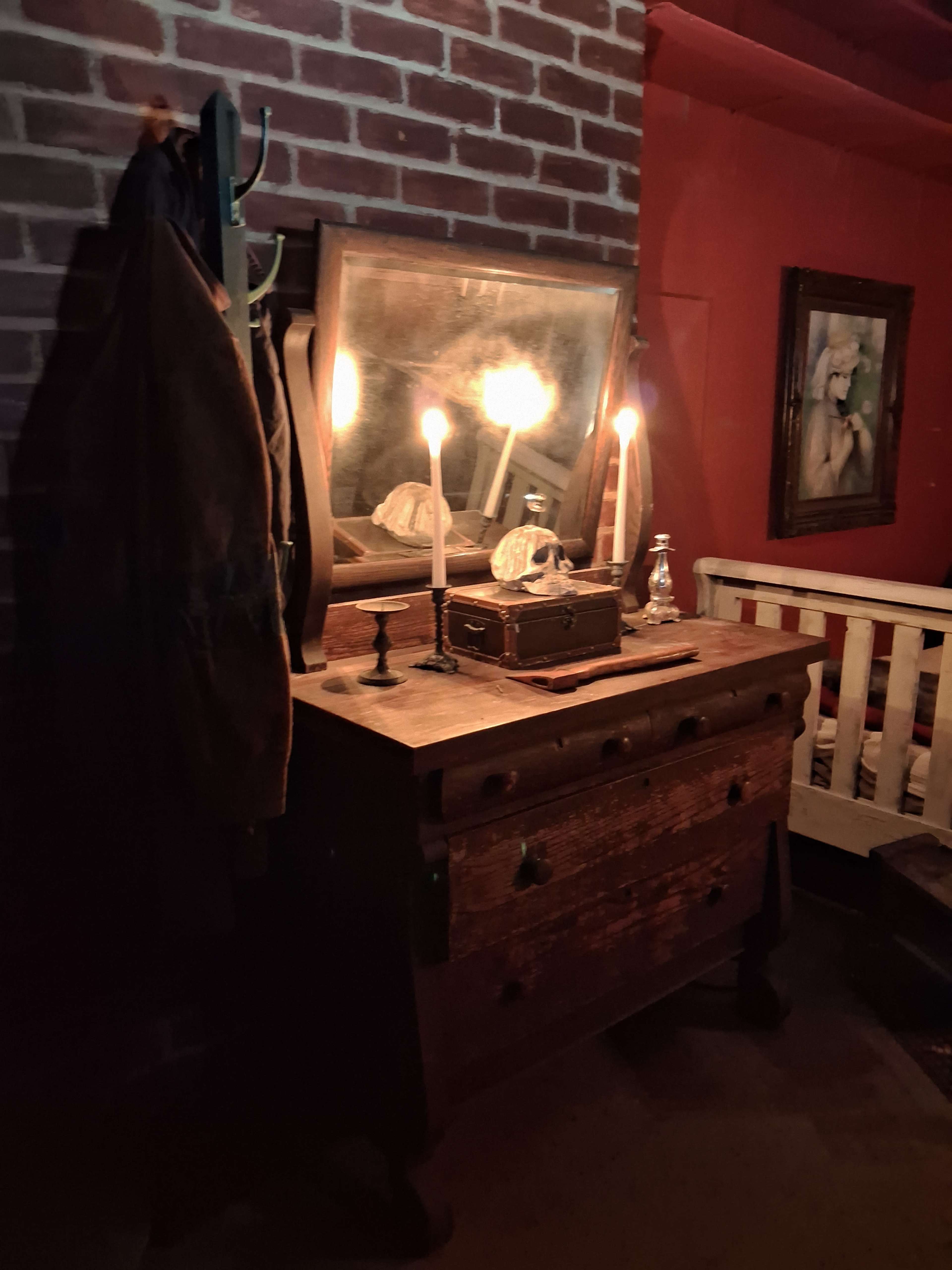 The scene features a wooden dresser with candles illuminating an antique mirror, against a backdrop of a red-brick wall and a framed painting.