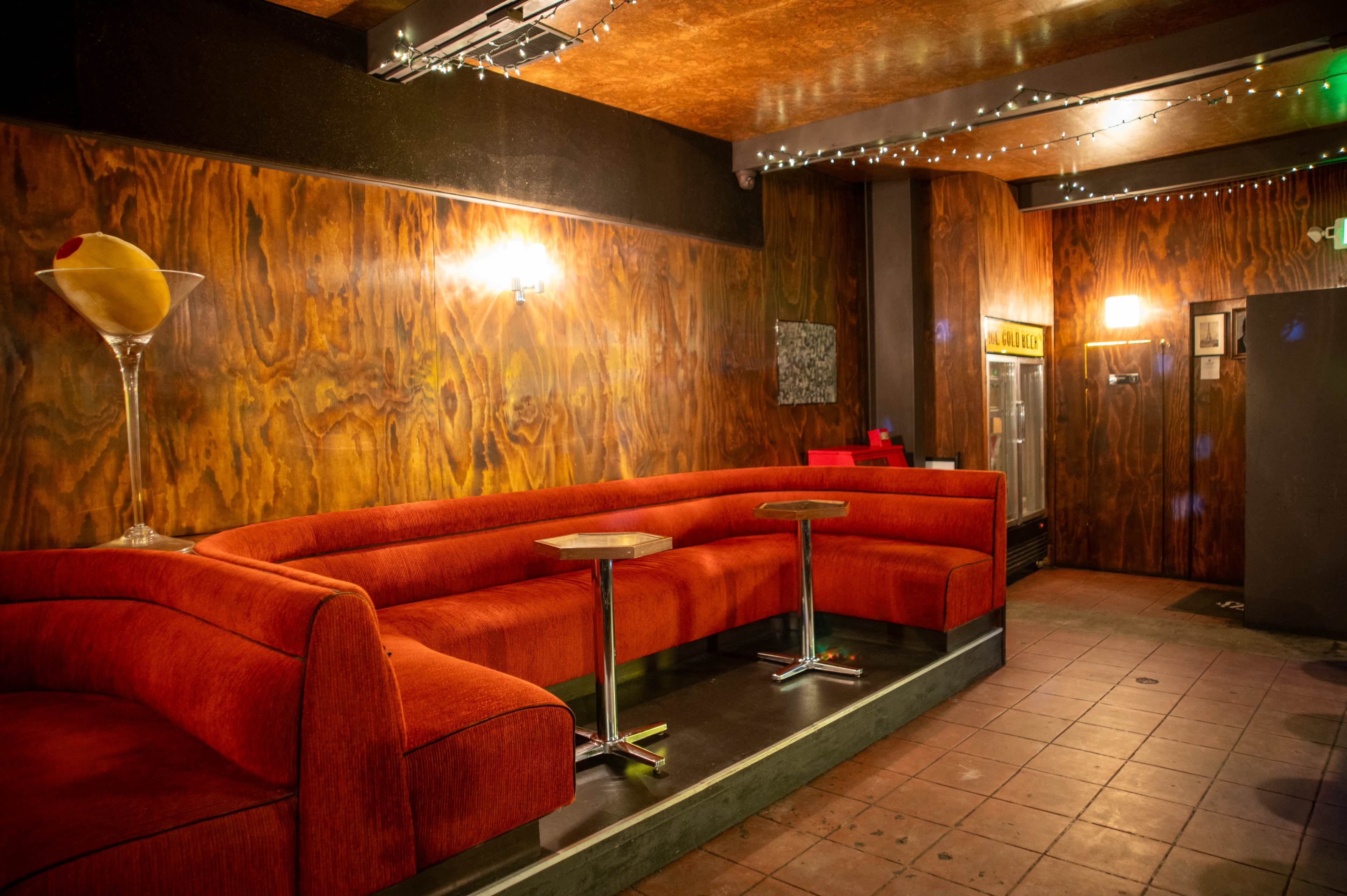 The image shows a cozy lounge area with orange curved sofas, small side tables, and dim lighting against a wooden wall backdrop.