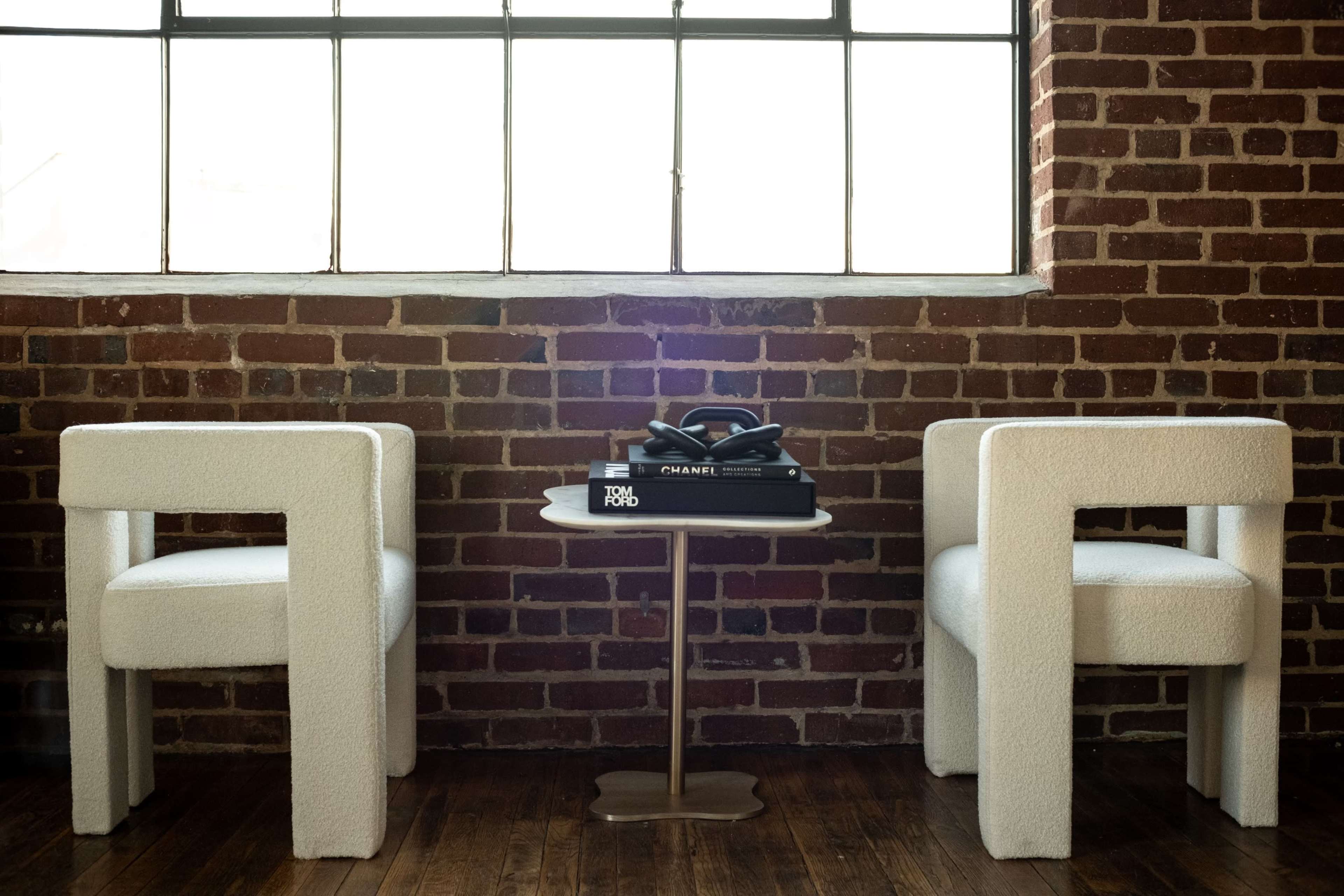 Two upholstered chairs frame a small round table, which holds a stack of books and a vintage telephone, against a backdrop of a brick wall and a large window.