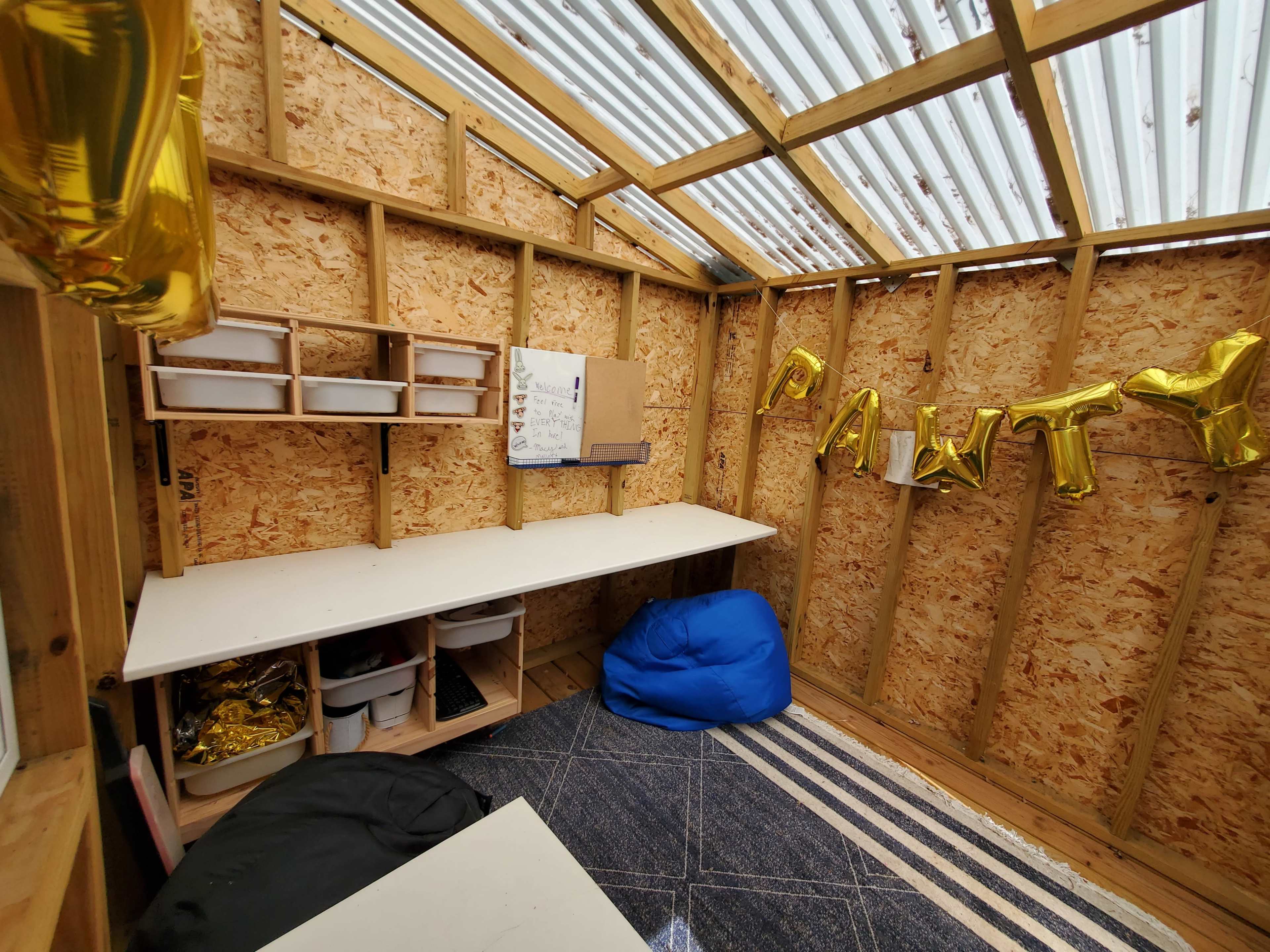 The interior of a wooden structure features a large table, storage shelves, and colorful balloons spelling "PARTY" along the wall.