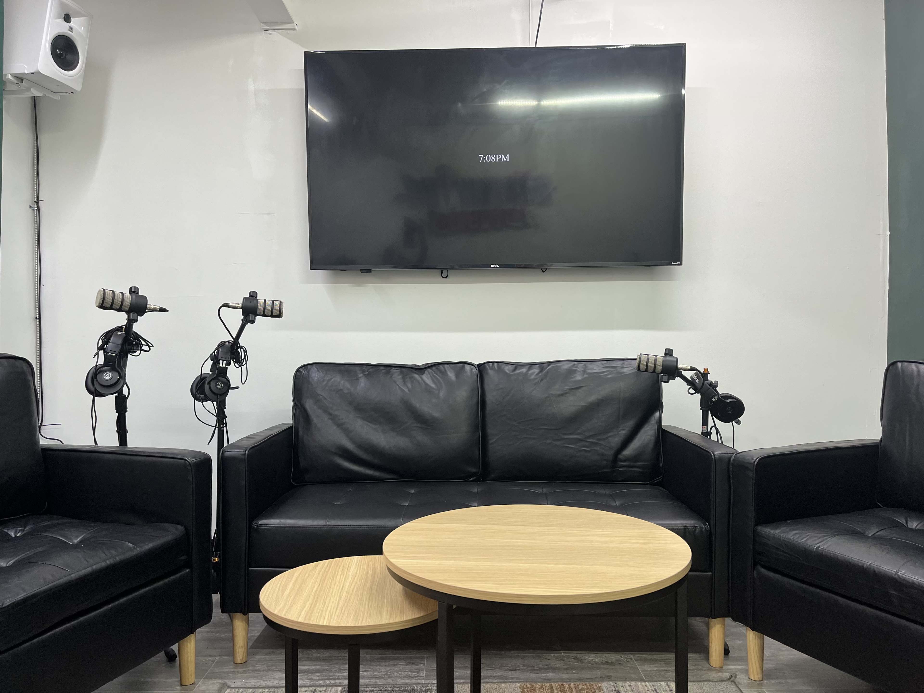 A small lounge area with two black leather sofas, a wall-mounted television, and two microphones on stands beside the seating.