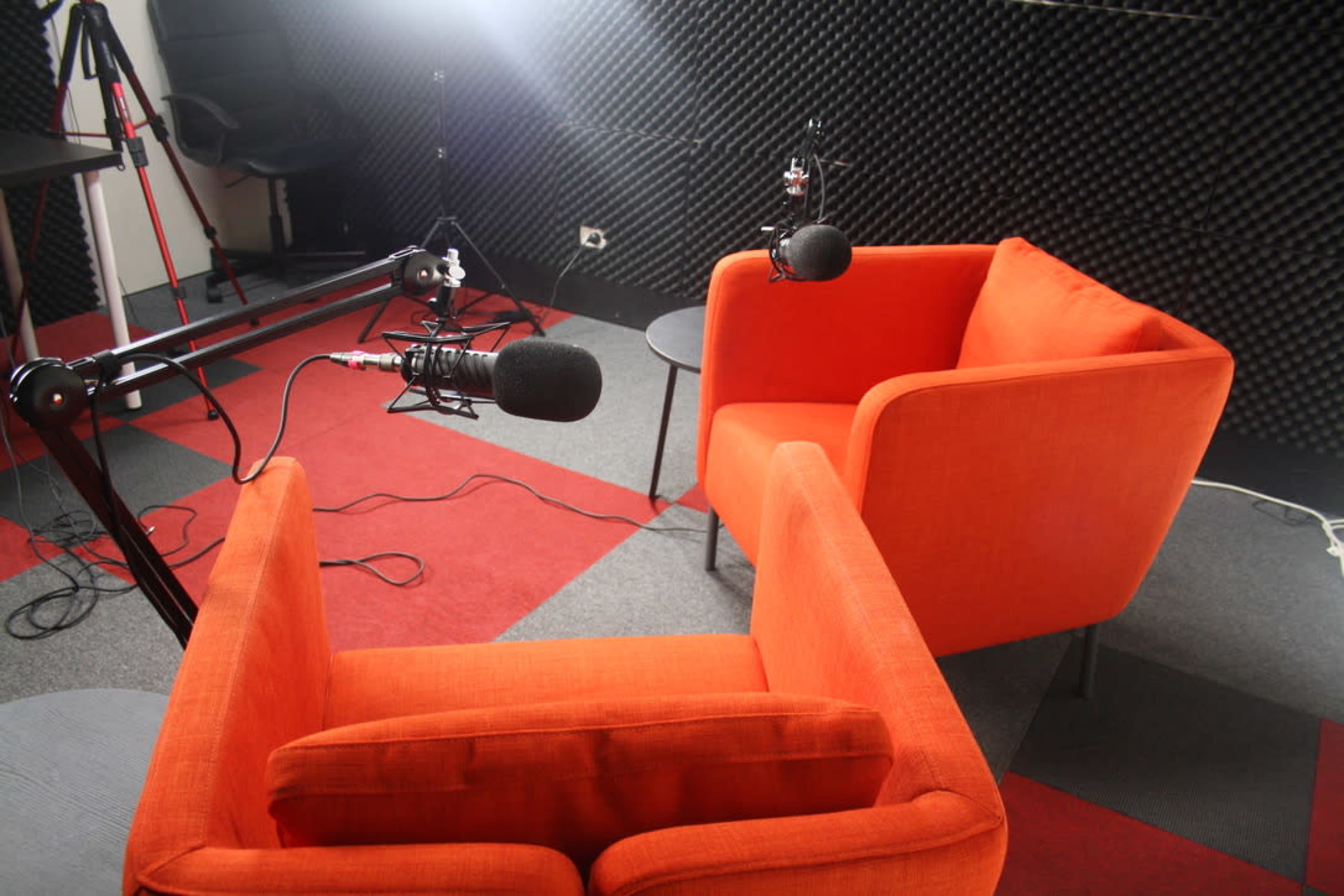 Two orange chairs facing each other in a studio setup with microphones and soundproofing materials in the background.