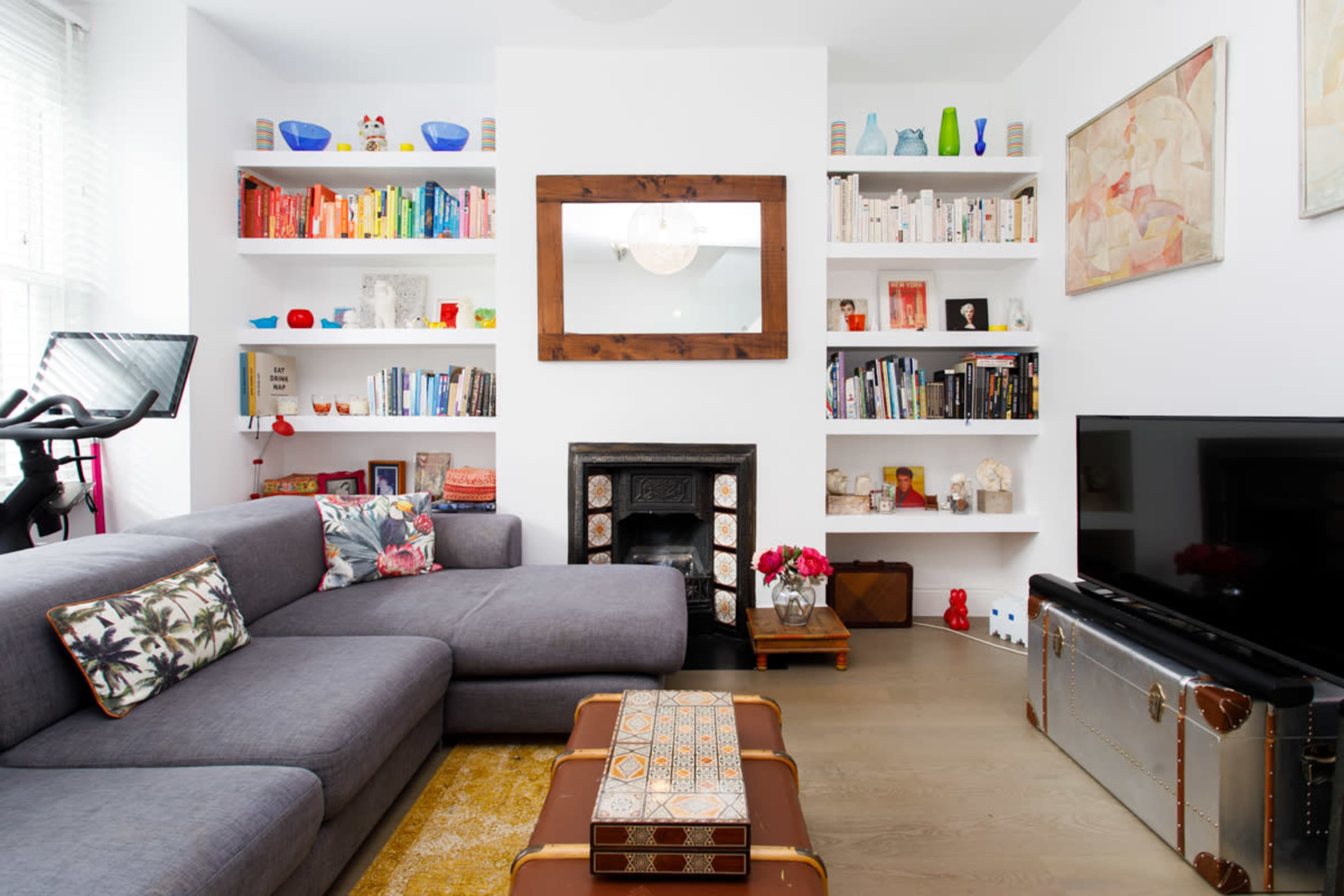 The image shows a modern living room featuring a gray sectional sofa, shelves filled with colorful books and decorative items, a flat-screen TV, and a coffee table.