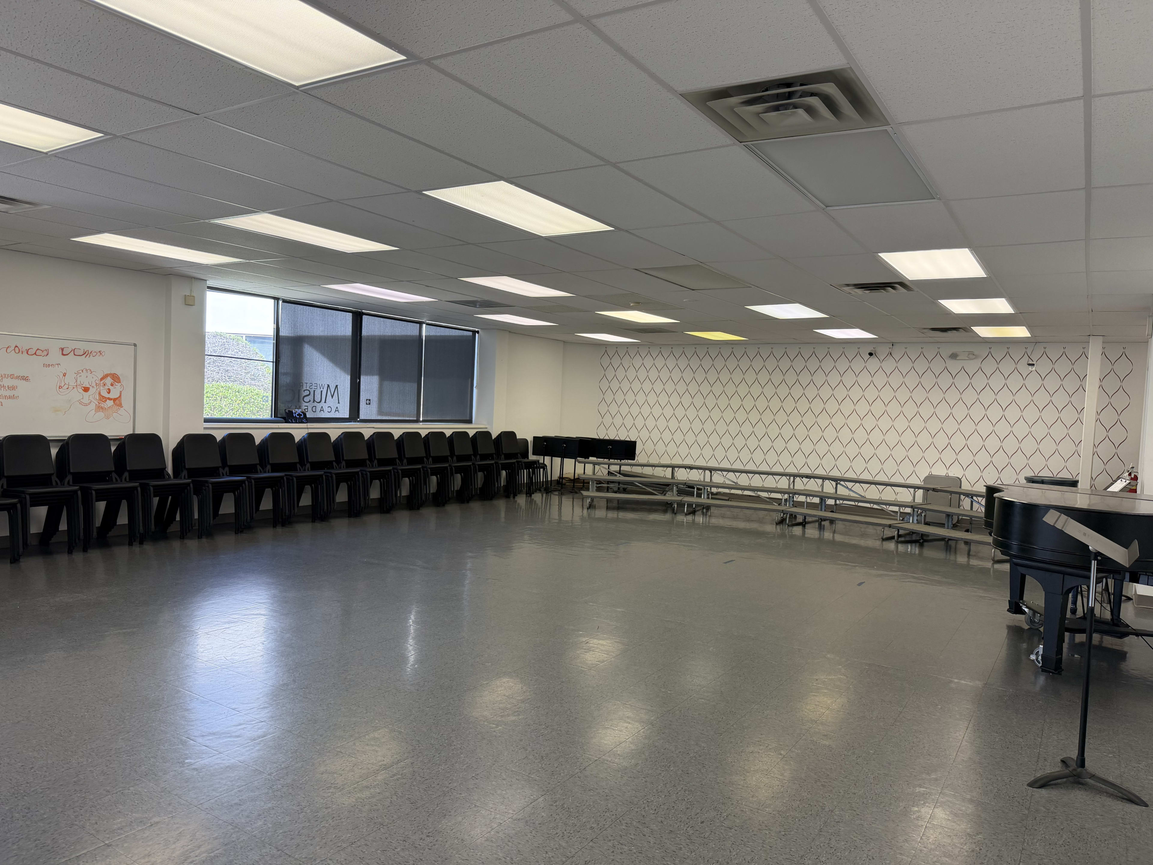 The image shows a spacious room with a row of black chairs along one wall, a set of risers in the center, and a piano positioned at the side.