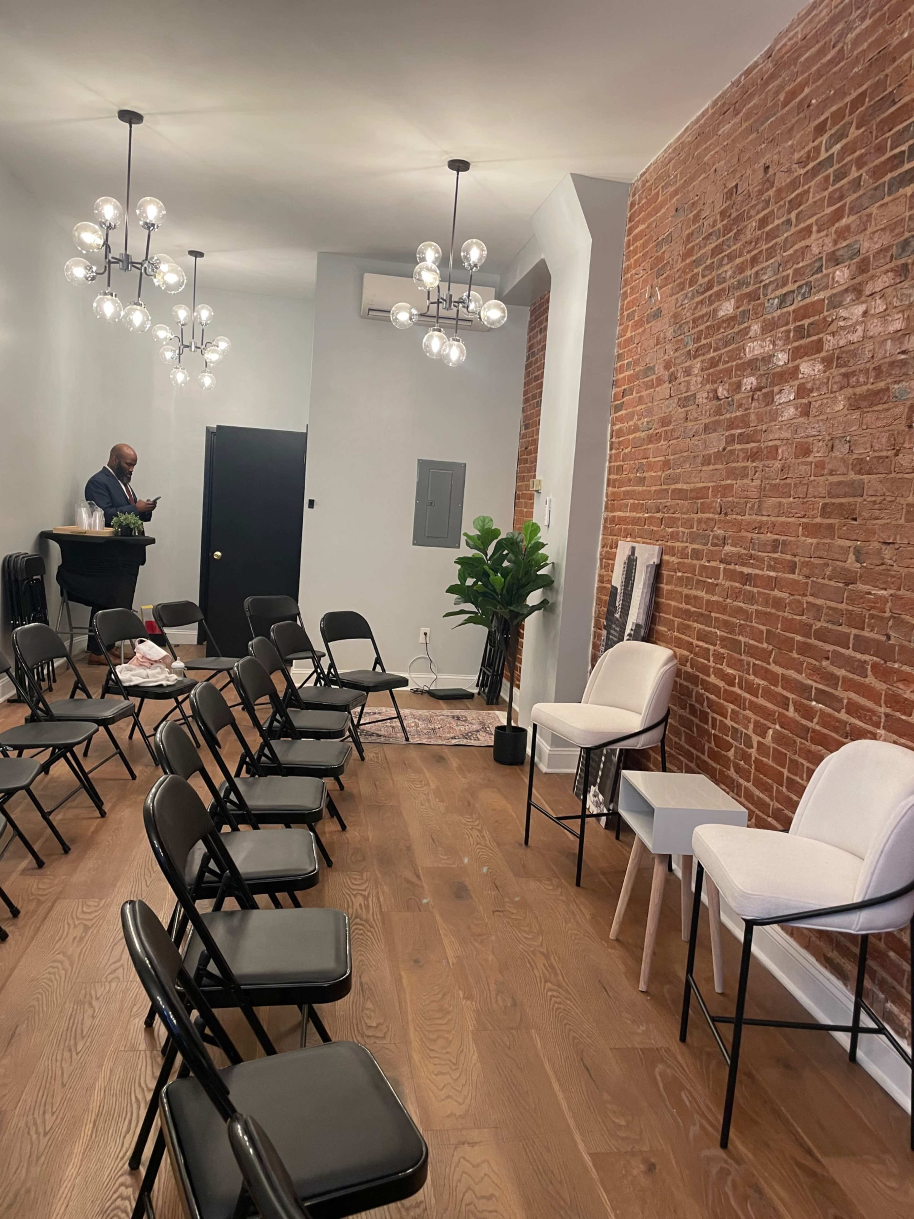 The image shows a room with several rows of black folding chairs facing a counter, adorned with a brick wall and modern lighting fixtures.