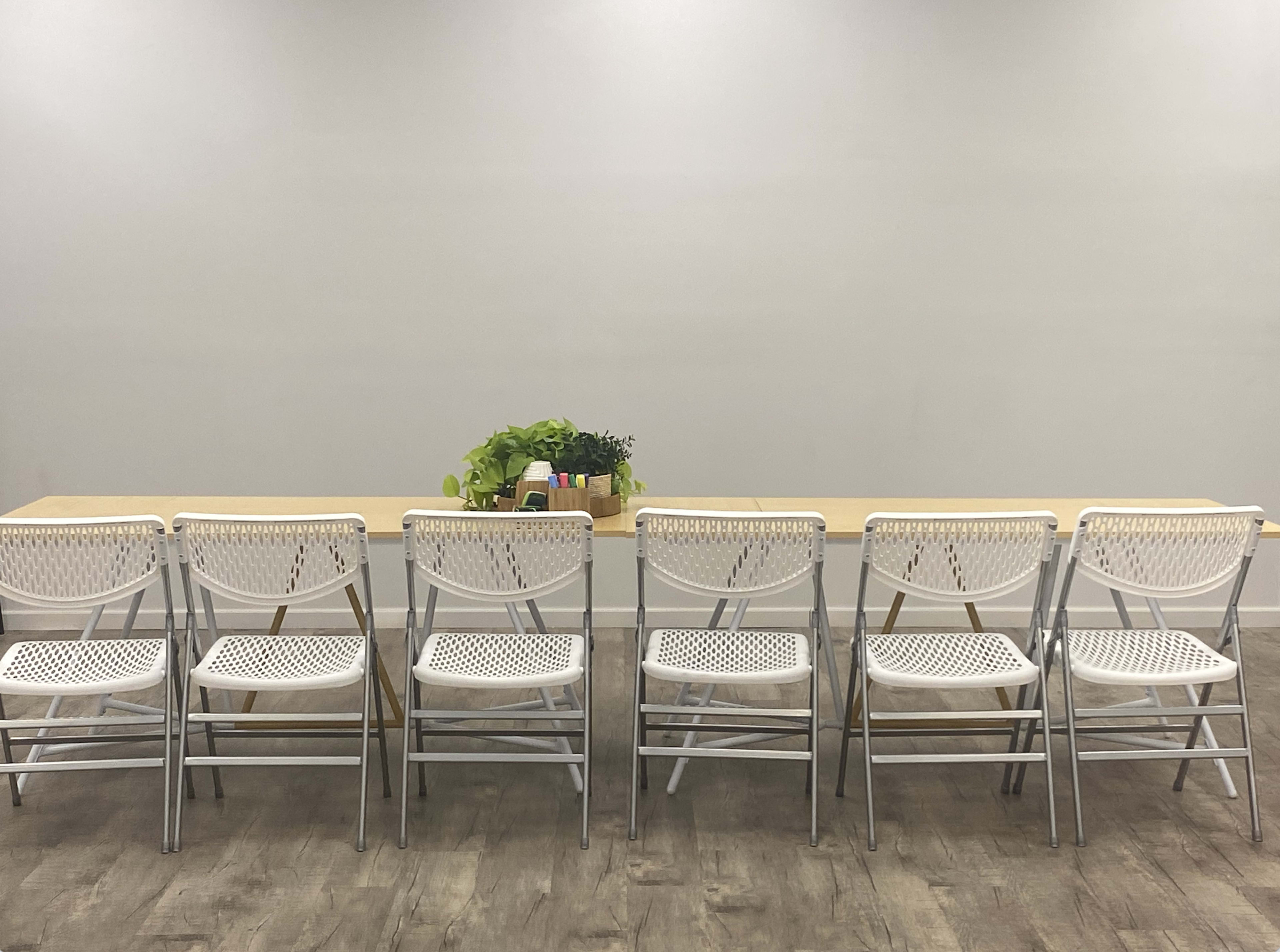 A row of six white folding chairs is lined up facing a long wooden table with a small potted plant on it.