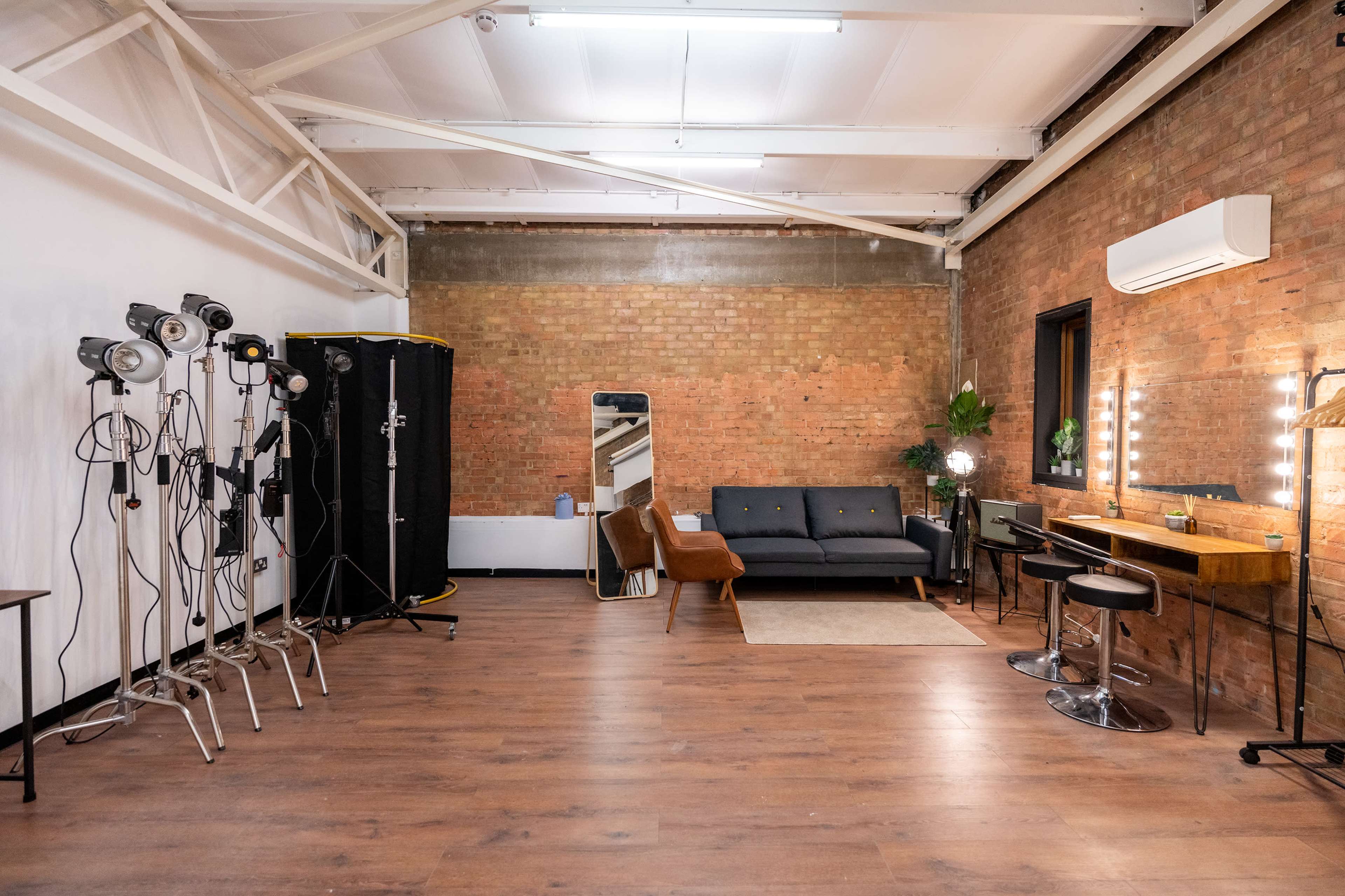 A spacious studio with wooden flooring, exposed brick walls, a black sofa, a mirror, and lighting equipment arranged on one side.