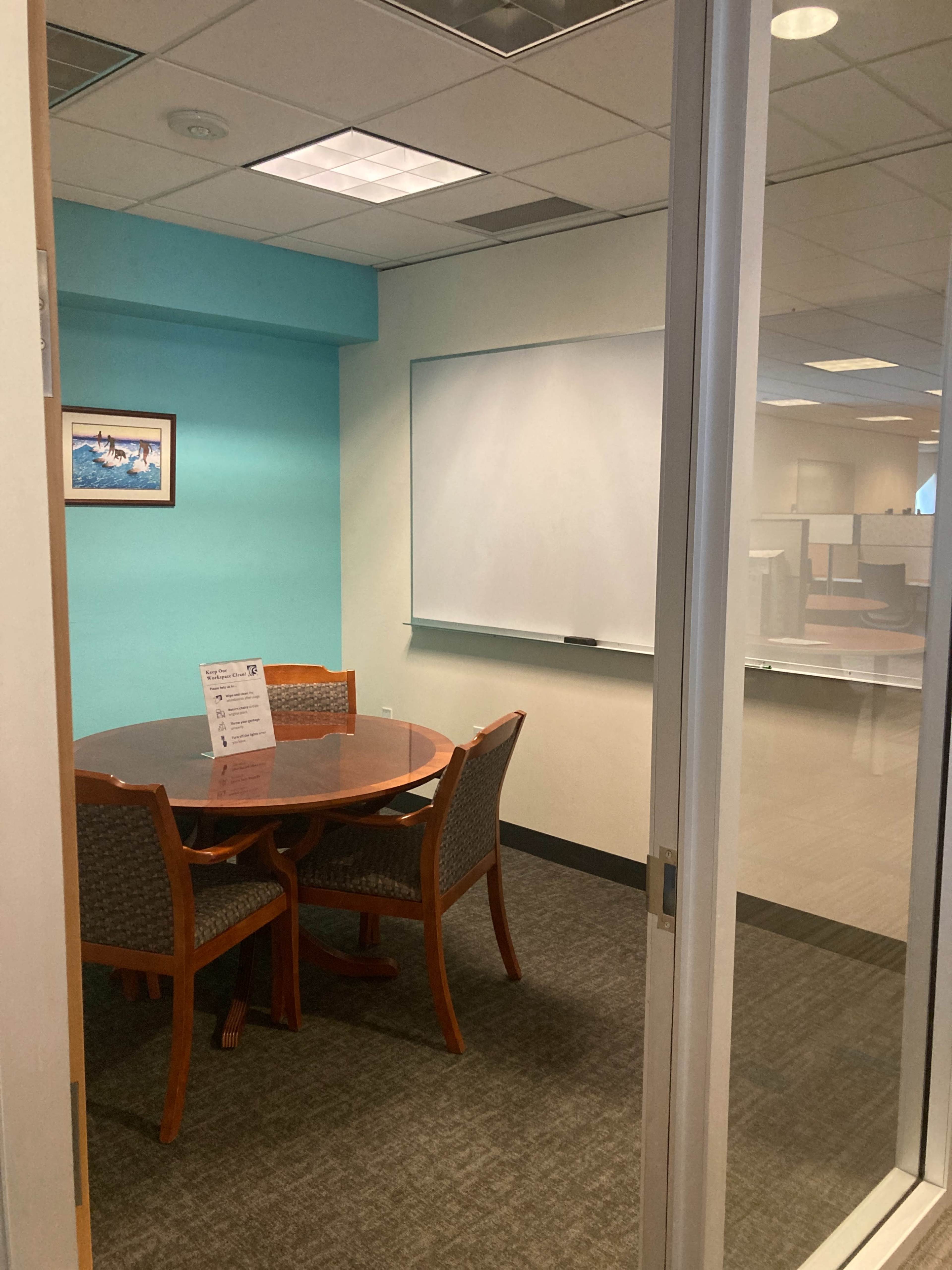 A small conference room featuring a round wooden table surrounded by four chairs, a whiteboard on the wall, and a turquoise-colored wall section.