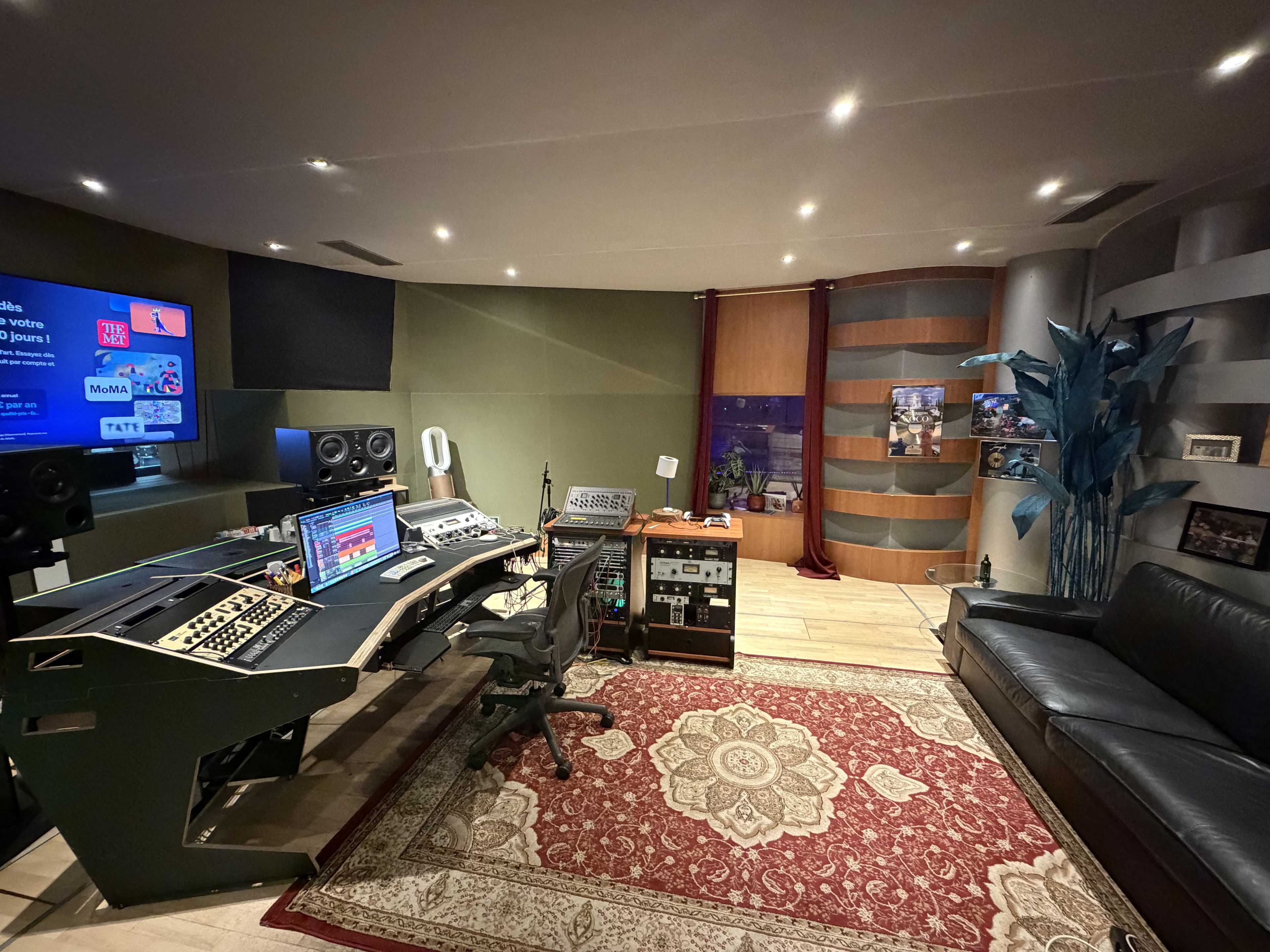 The image shows a modern music recording studio featuring a mixing console, multiple monitors, sound equipment, and a comfortable seating area with a patterned rug.