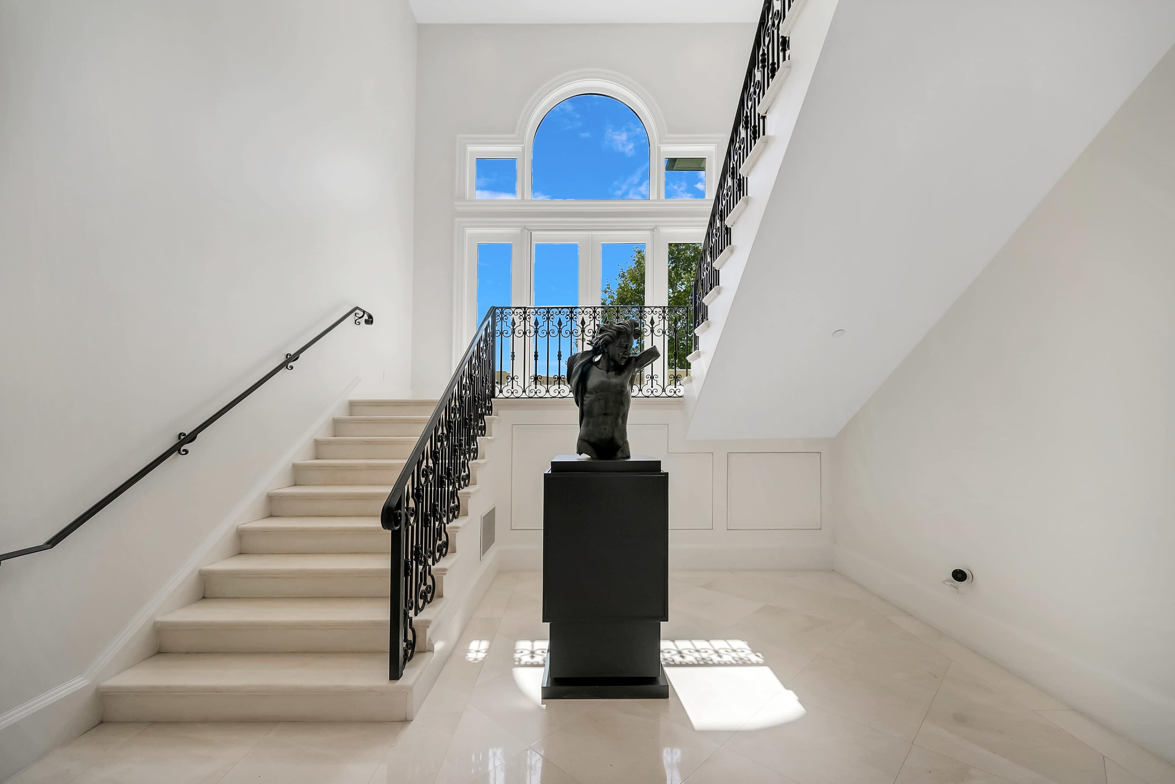 A staircase leads up from a bright foyer, featuring a statue on a pedestal beneath a large arched window.