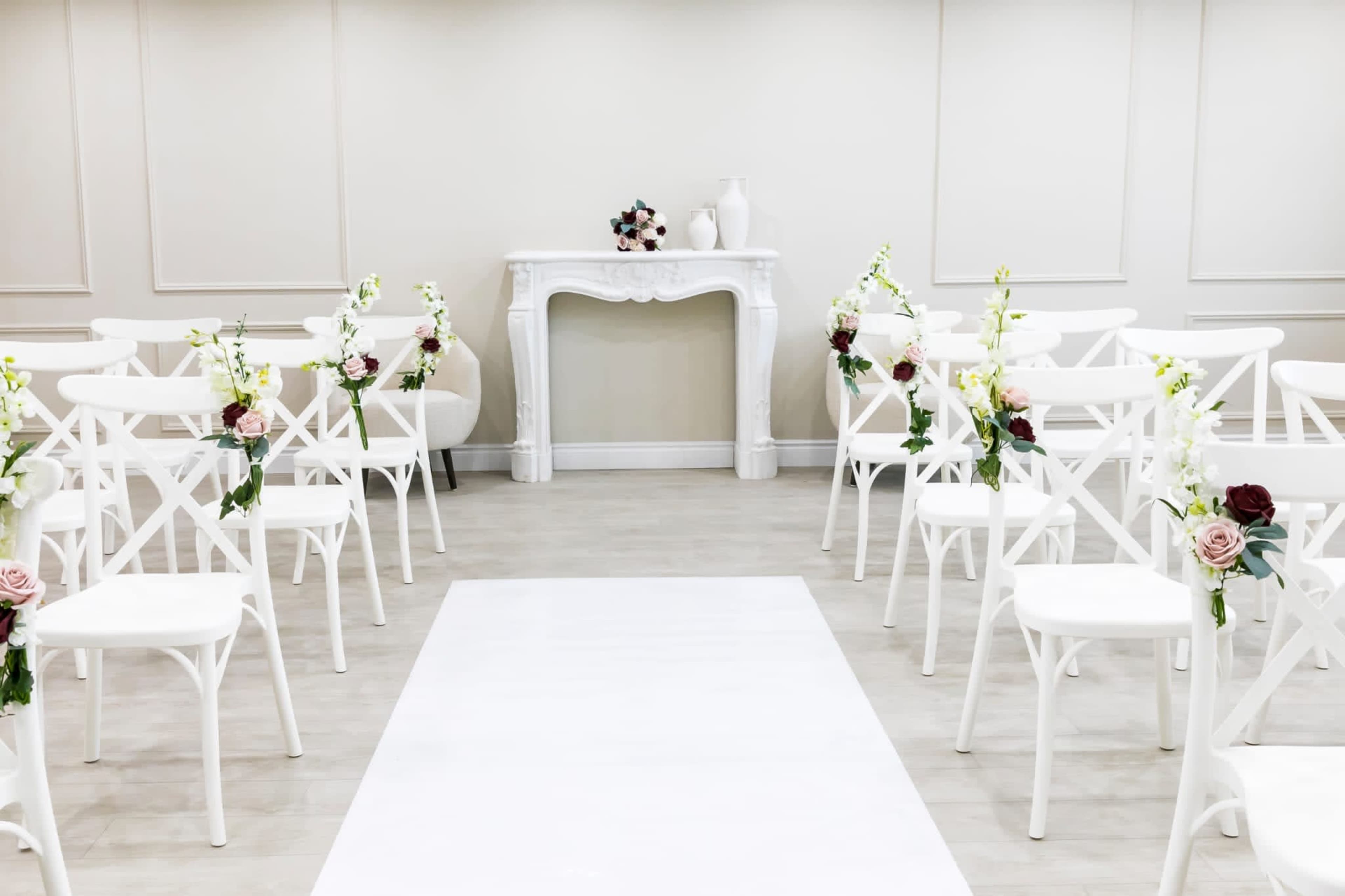 The scene features a minimalist wedding ceremony setup with white chairs arranged in rows facing a central white aisle and a decorative mantelpiece adorned with flowers.