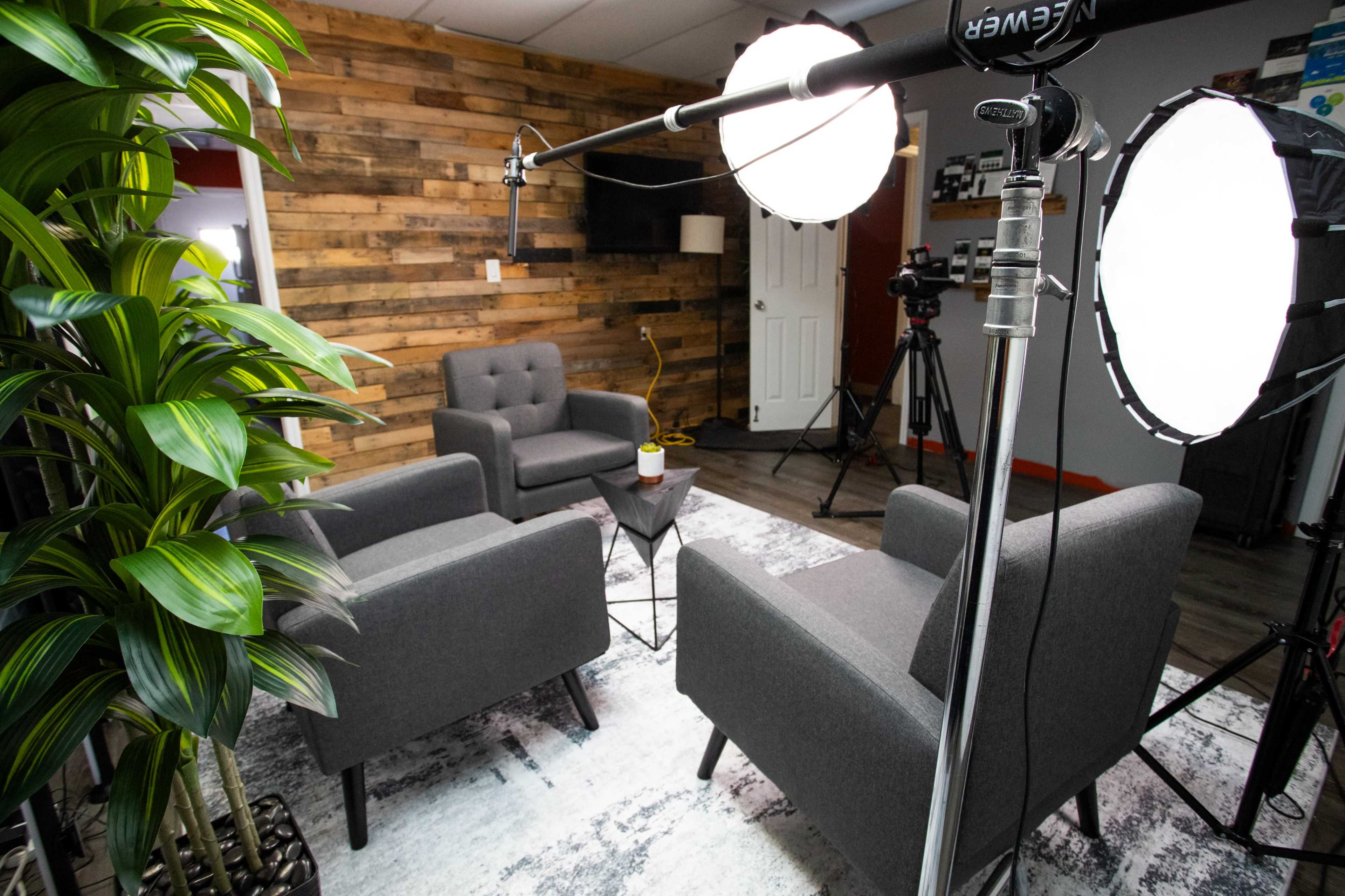 The image shows a well-lit studio setup featuring two gray armchairs facing a small black table on a patterned rug, with a backdrop of wooden panels and camera equipment in the background.