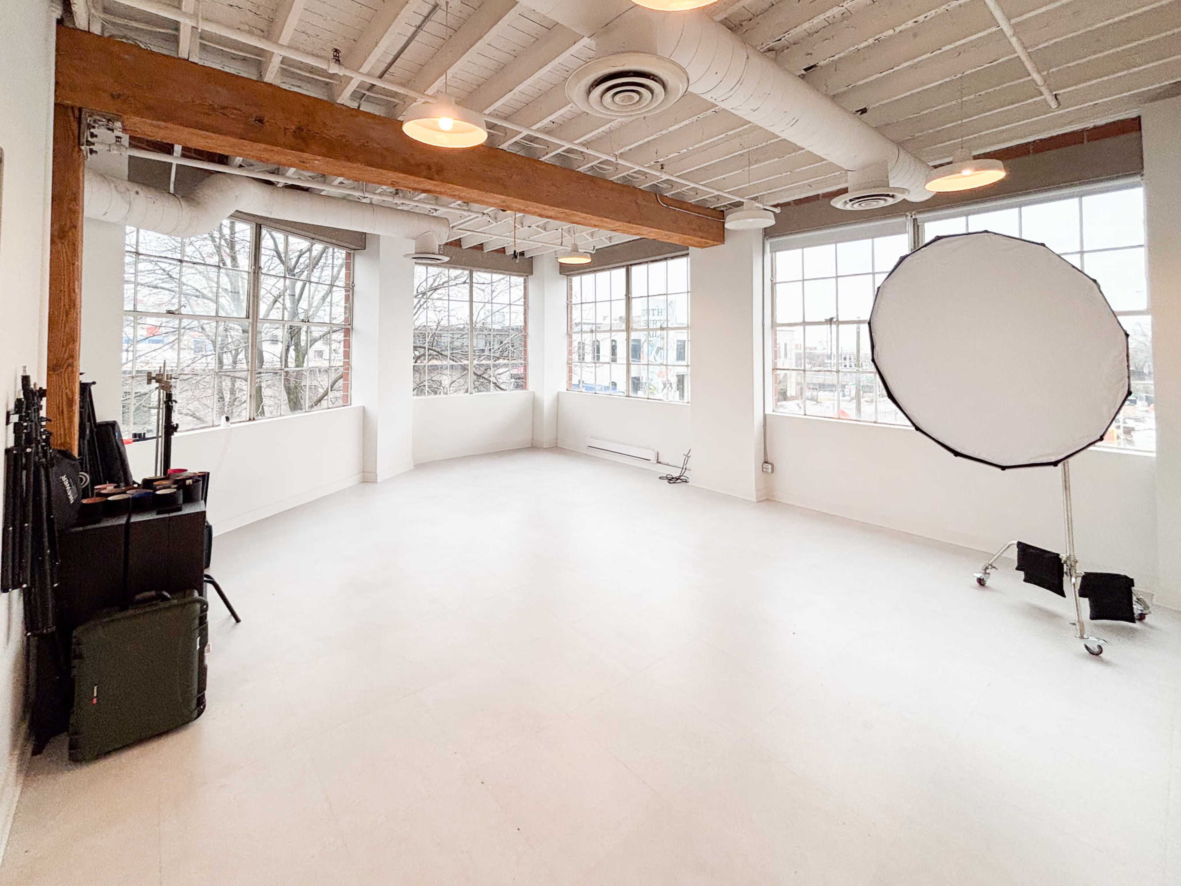 The image shows an empty studio space with large windows, wooden beams, and photography equipment in the corner.