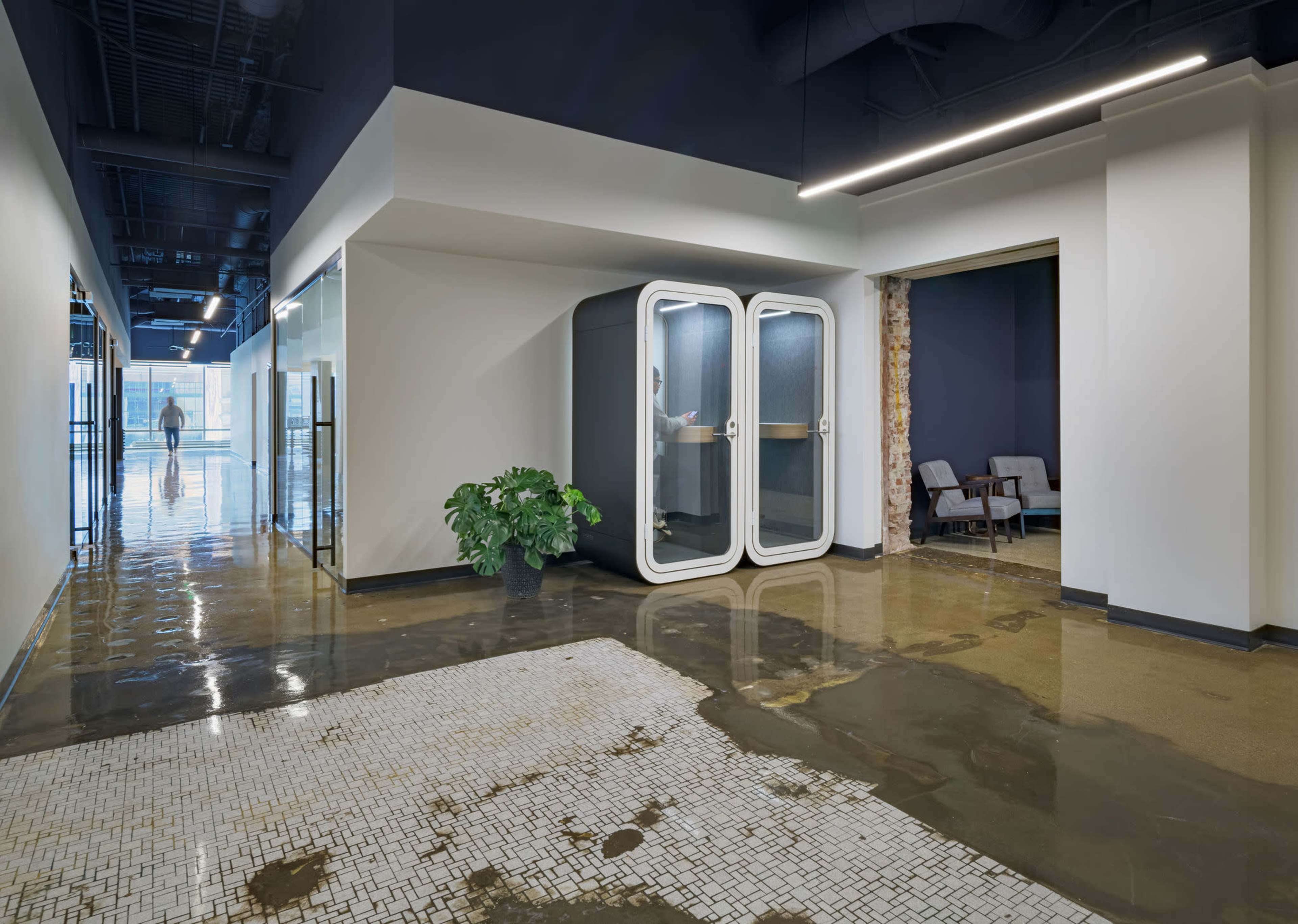 The image shows a modern office space with polished concrete flooring, a glass meeting pod, and an area with chairs behind a brick wall.