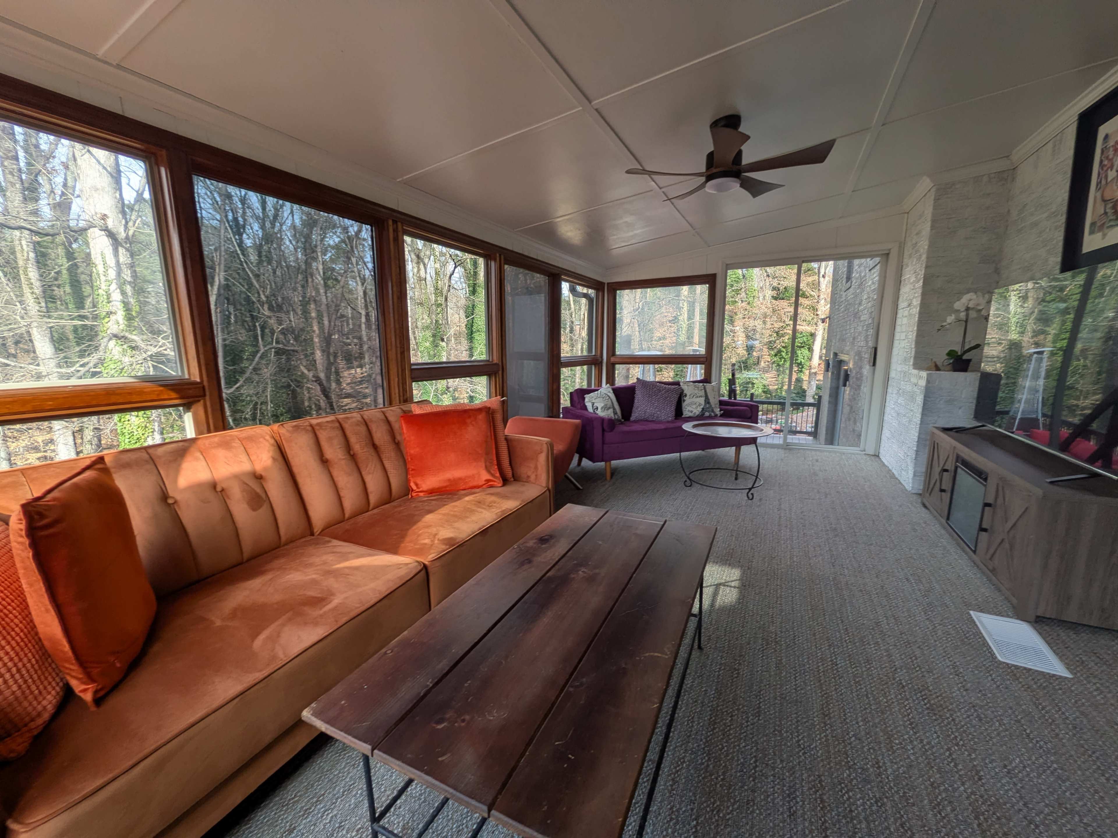 The image shows a screened-in porch with an orange sectional sofa, a purple armchair, a wooden coffee table, and large windows overlooking a wooded area.