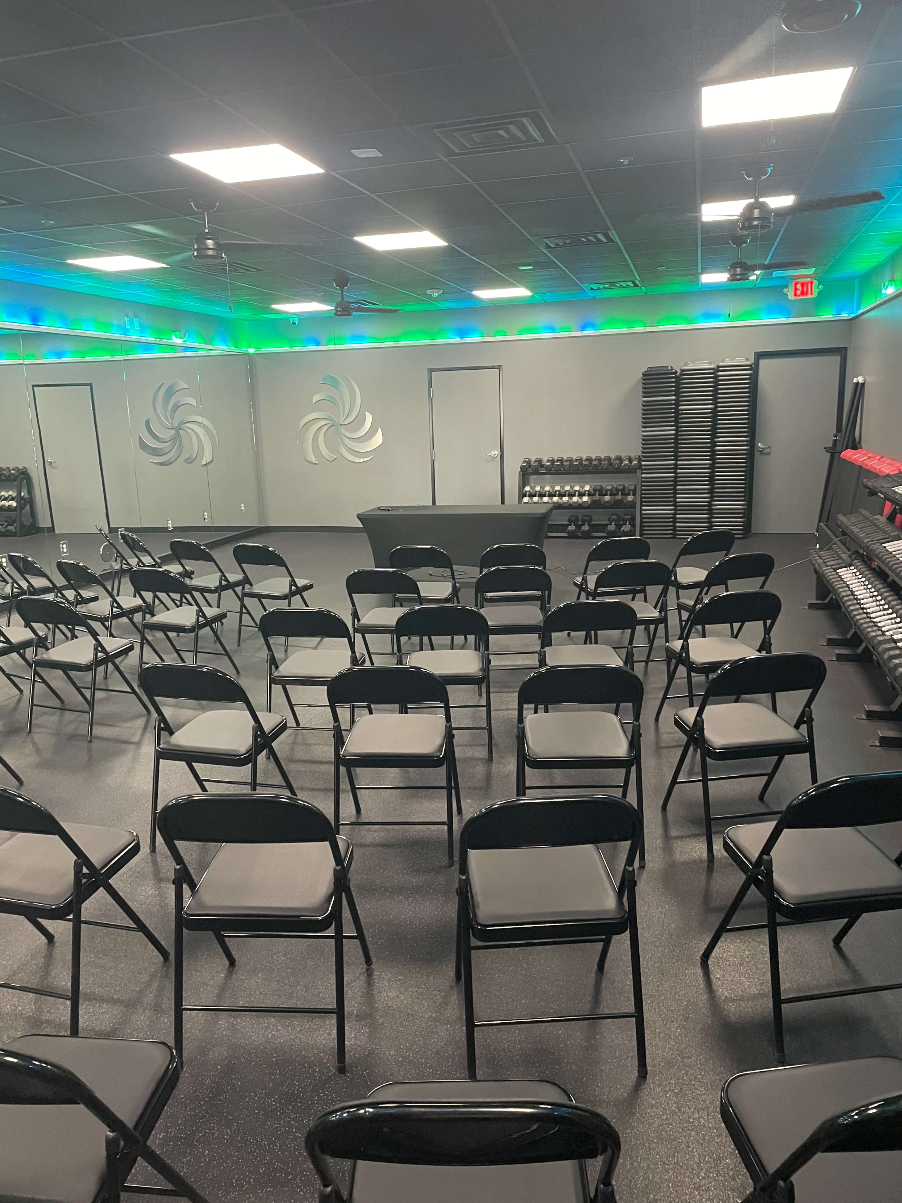 The image shows a fitness studio with rows of black folding chairs arranged facing a table, mirrored walls, and shelves of free weights in the background.