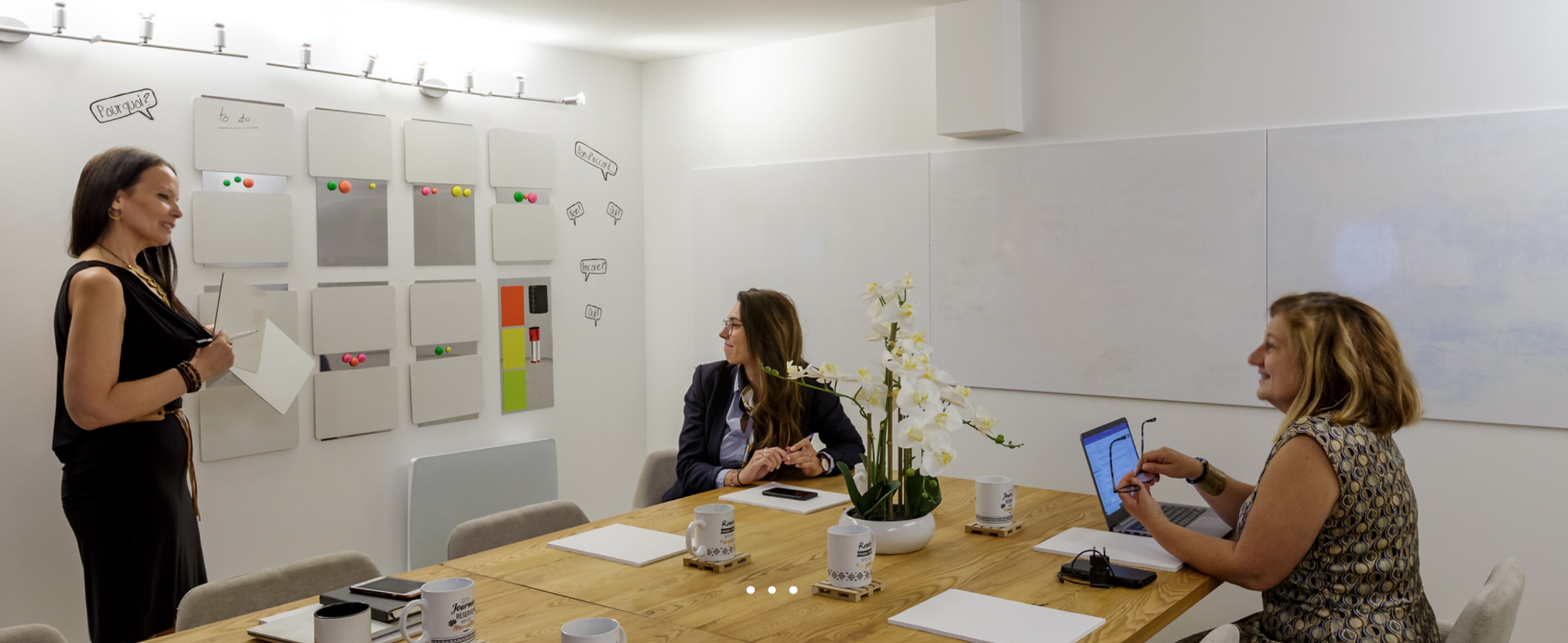 A woman presents in a meeting room while two others listen, with a wall displaying various colored sticky notes and a table set with mugs and notepads.