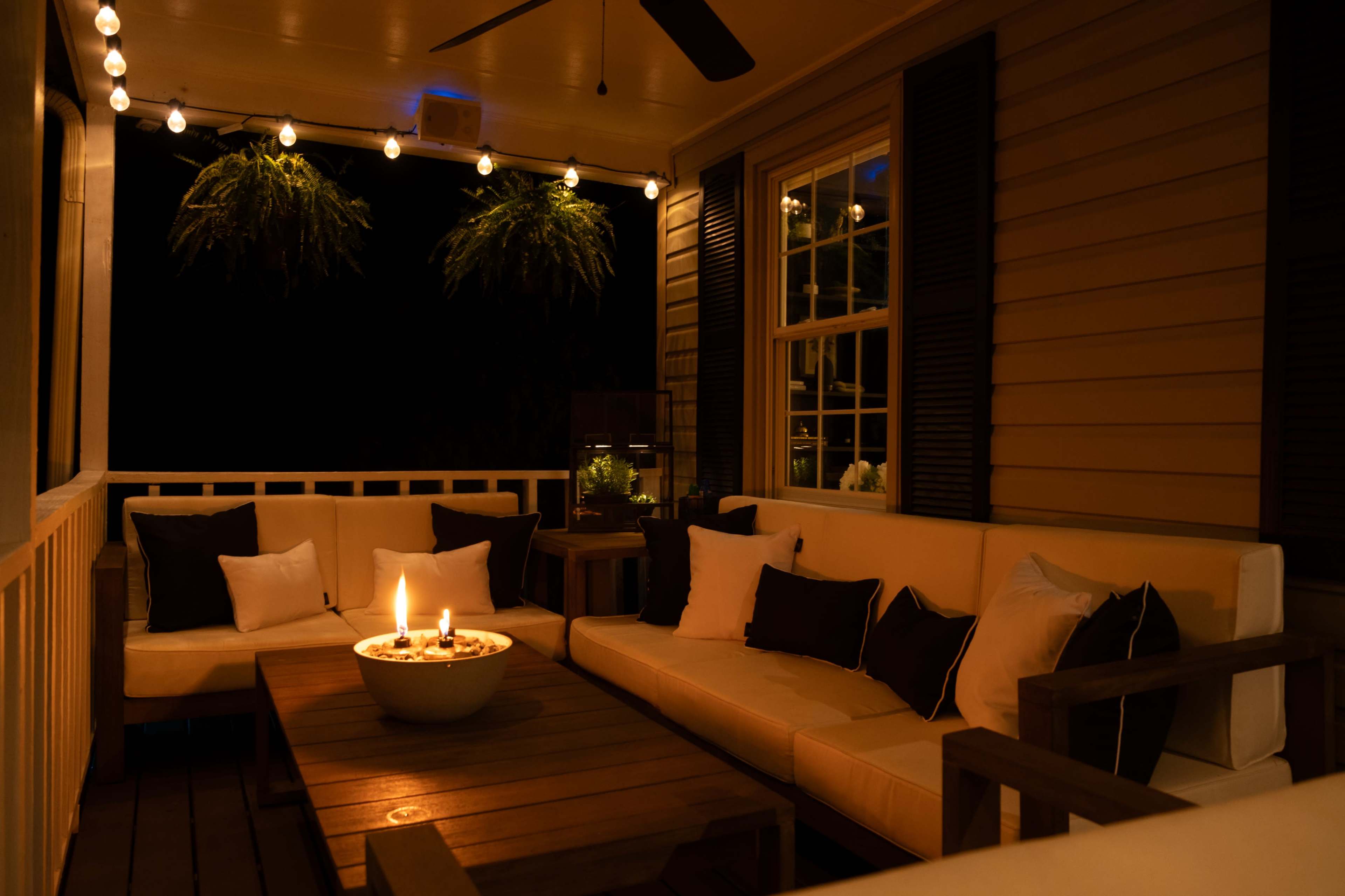 A cozy porch area features a seating arrangement with a fire pit at the center, surrounded by string lights and lush greenery.