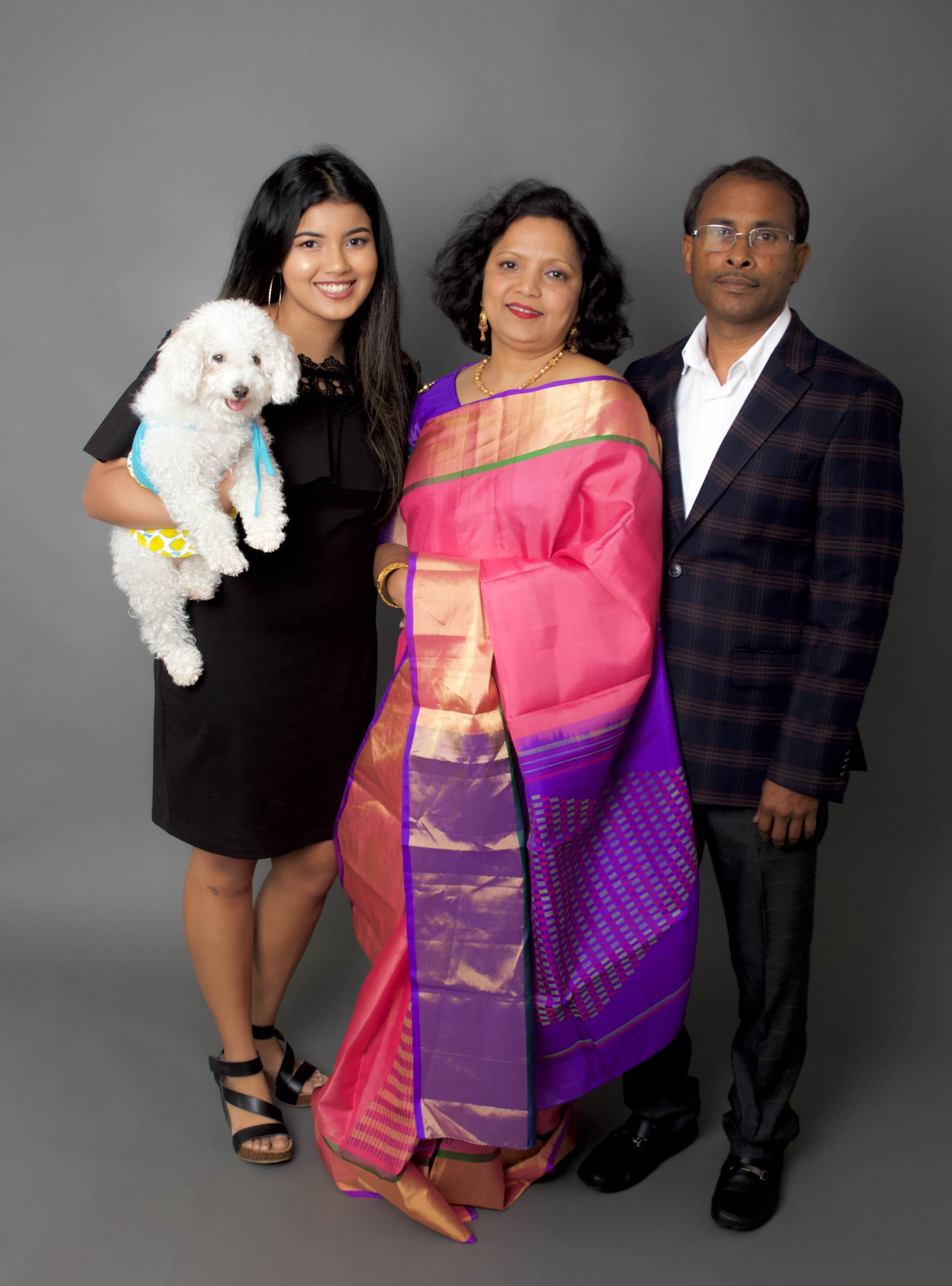 A woman in a black dress holds a small dog while standing next to a woman in a colorful sari and a man in a checkered suit.