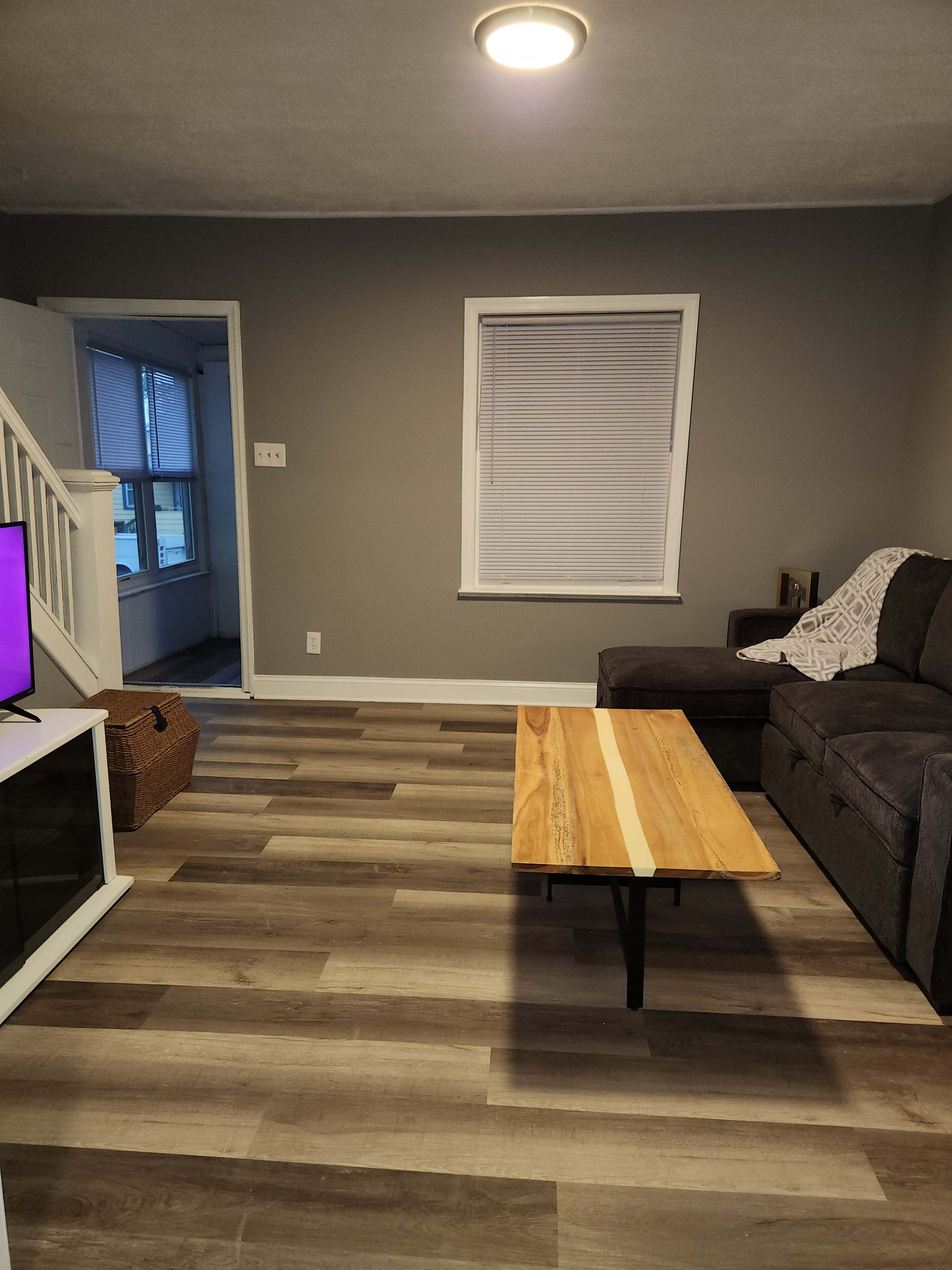 The image shows a living room with a gray wall, a dark sofa, a wooden coffee table, and a window allowing natural light.