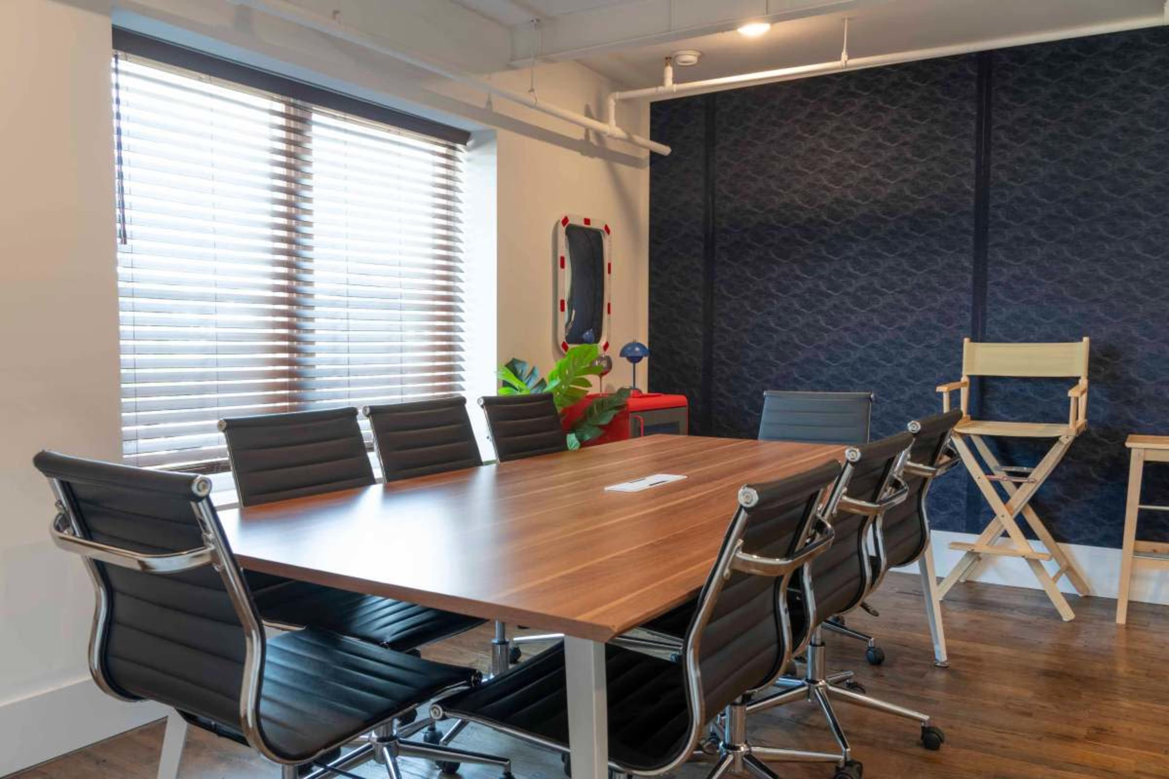 A conference room features a large wooden table surrounded by black ergonomic chairs, with blinds covering the windows and a dark patterned wall in the background.