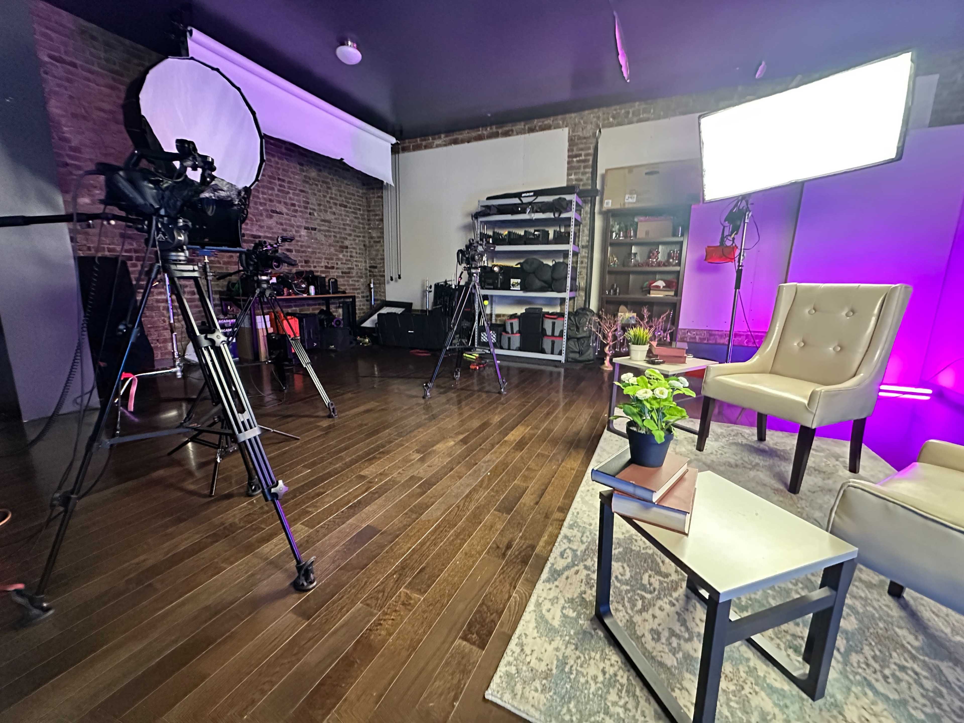 A television studio setup features cameras, lighting equipment, and a stylish seating area with plants and books on a side table.