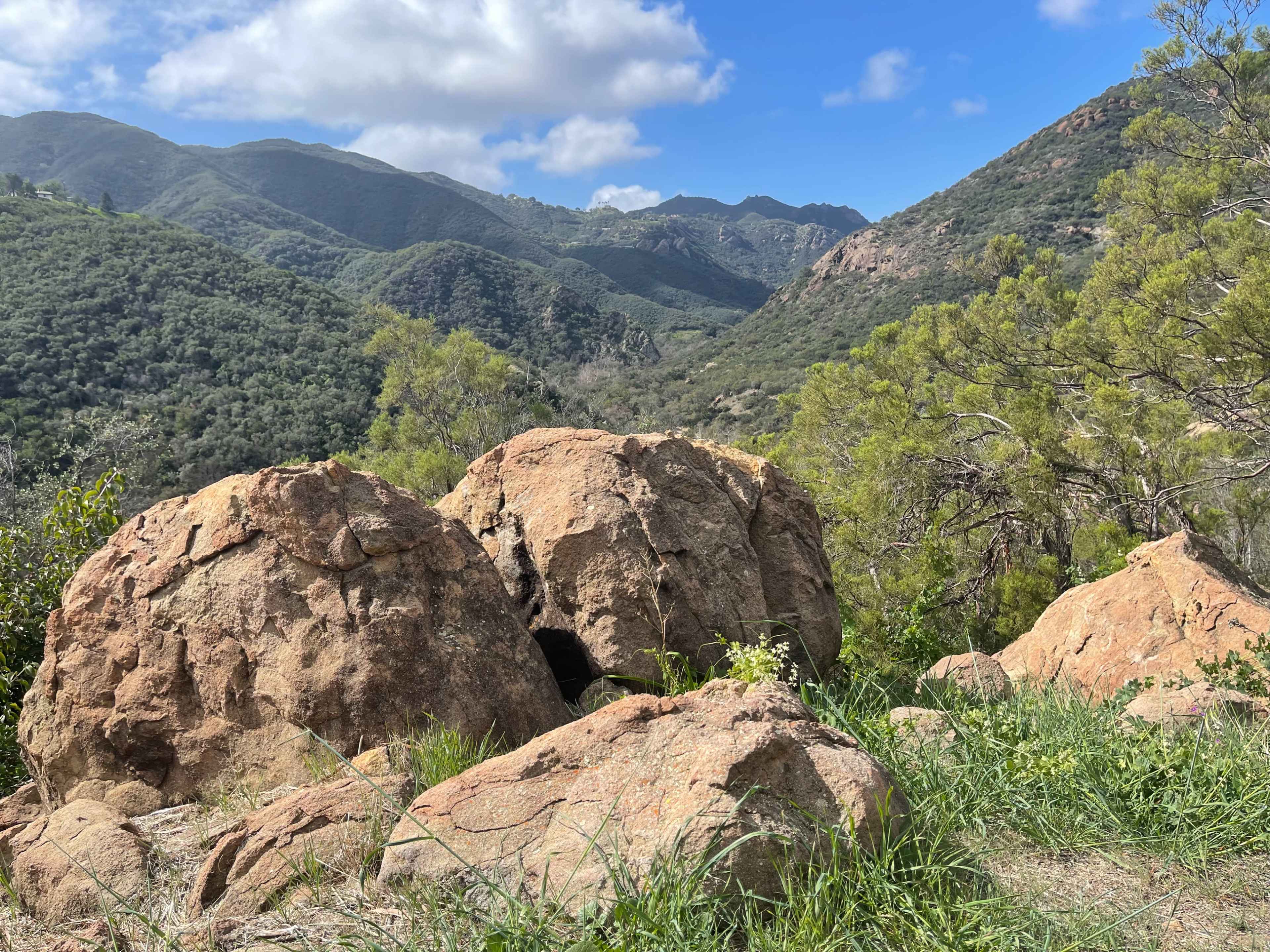 A cluster of large boulders sits at the forefront with rolling green hills and mountains in the background under a partly cloudy sky.