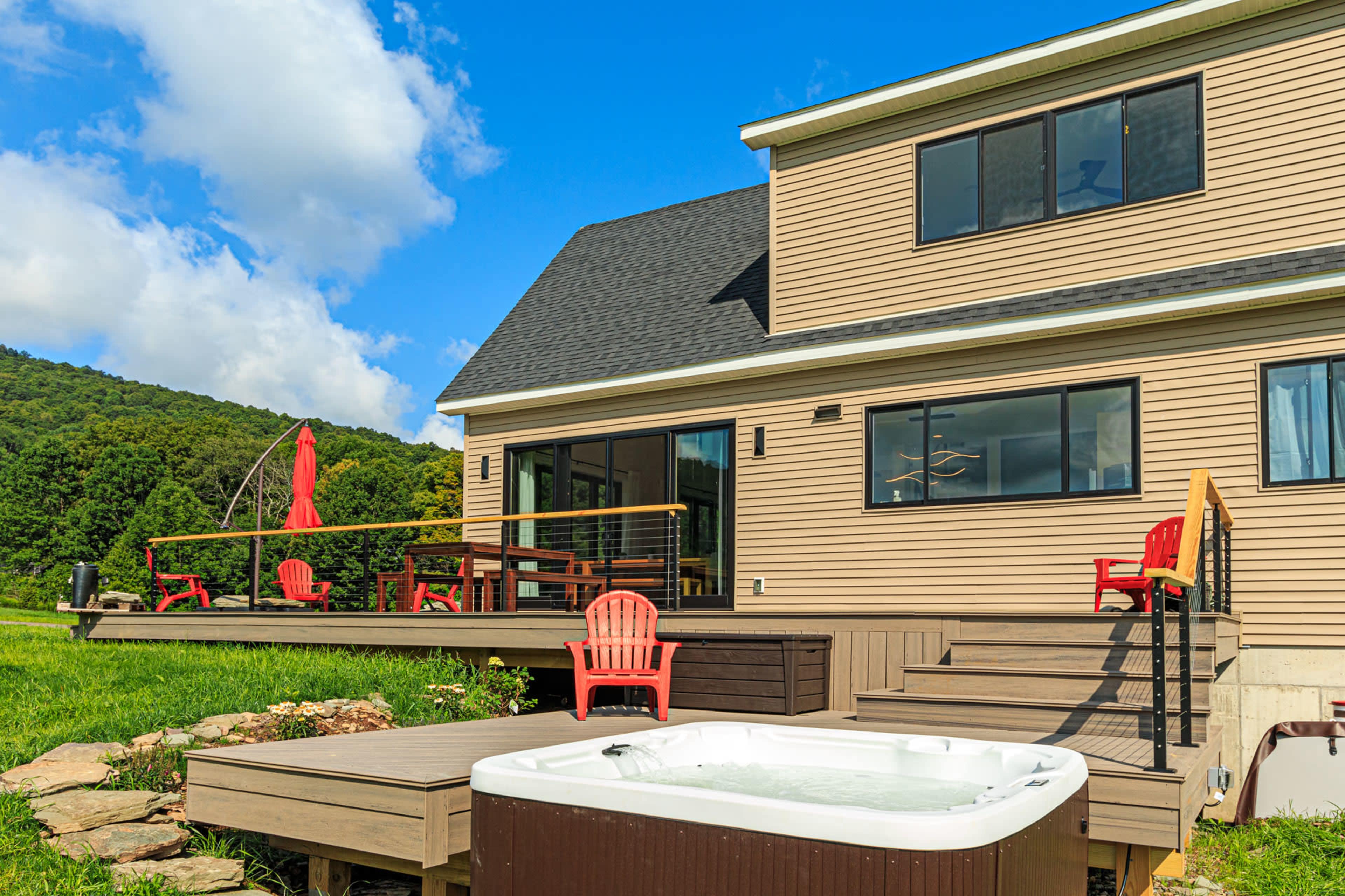 A house with a deck overlooks a green landscape, featuring red chairs and a hot tub in the foreground.