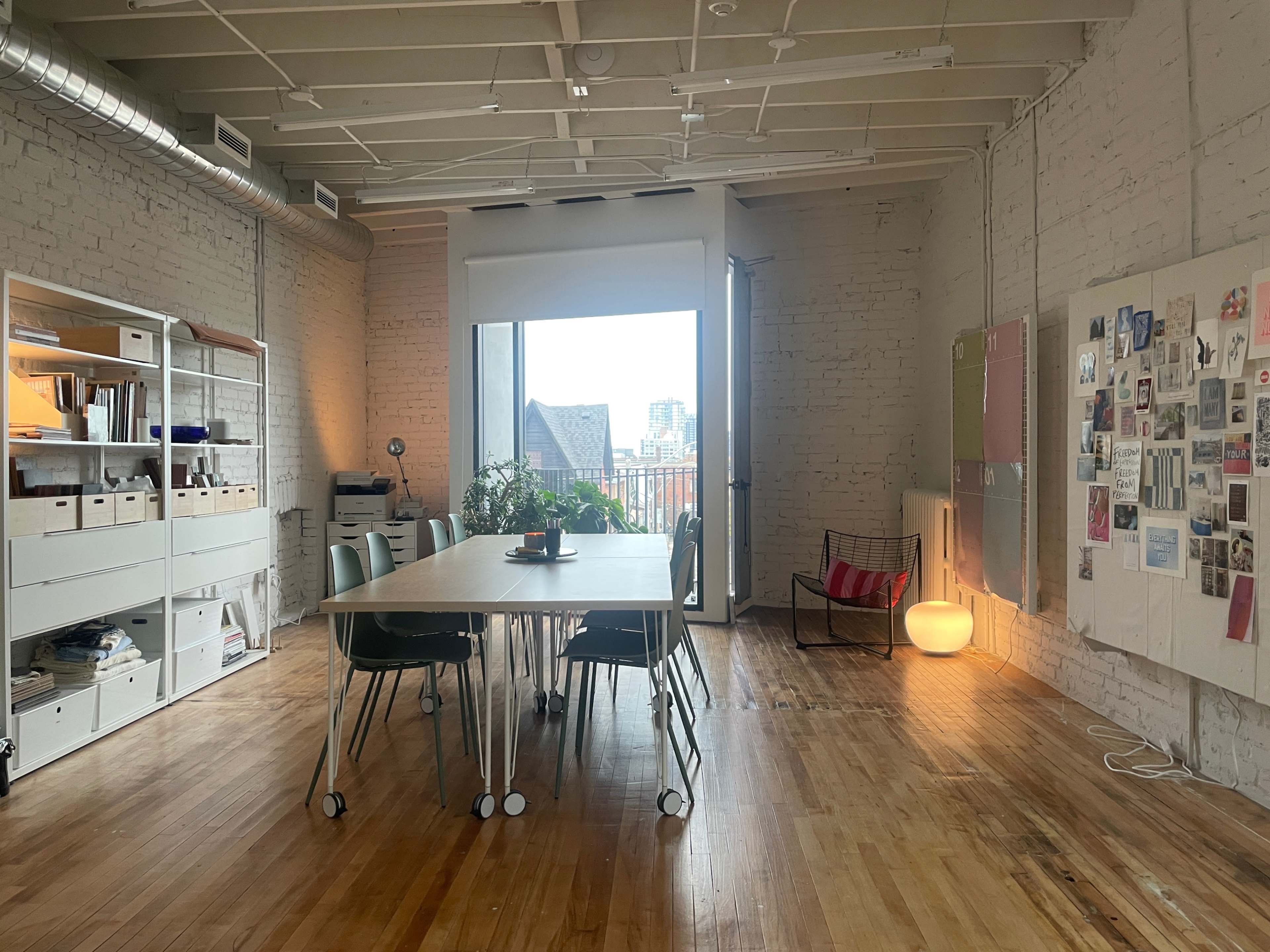A large table is surrounded by chairs in a sunny, minimalist room featuring exposed brick walls and a window showing a city skyline.