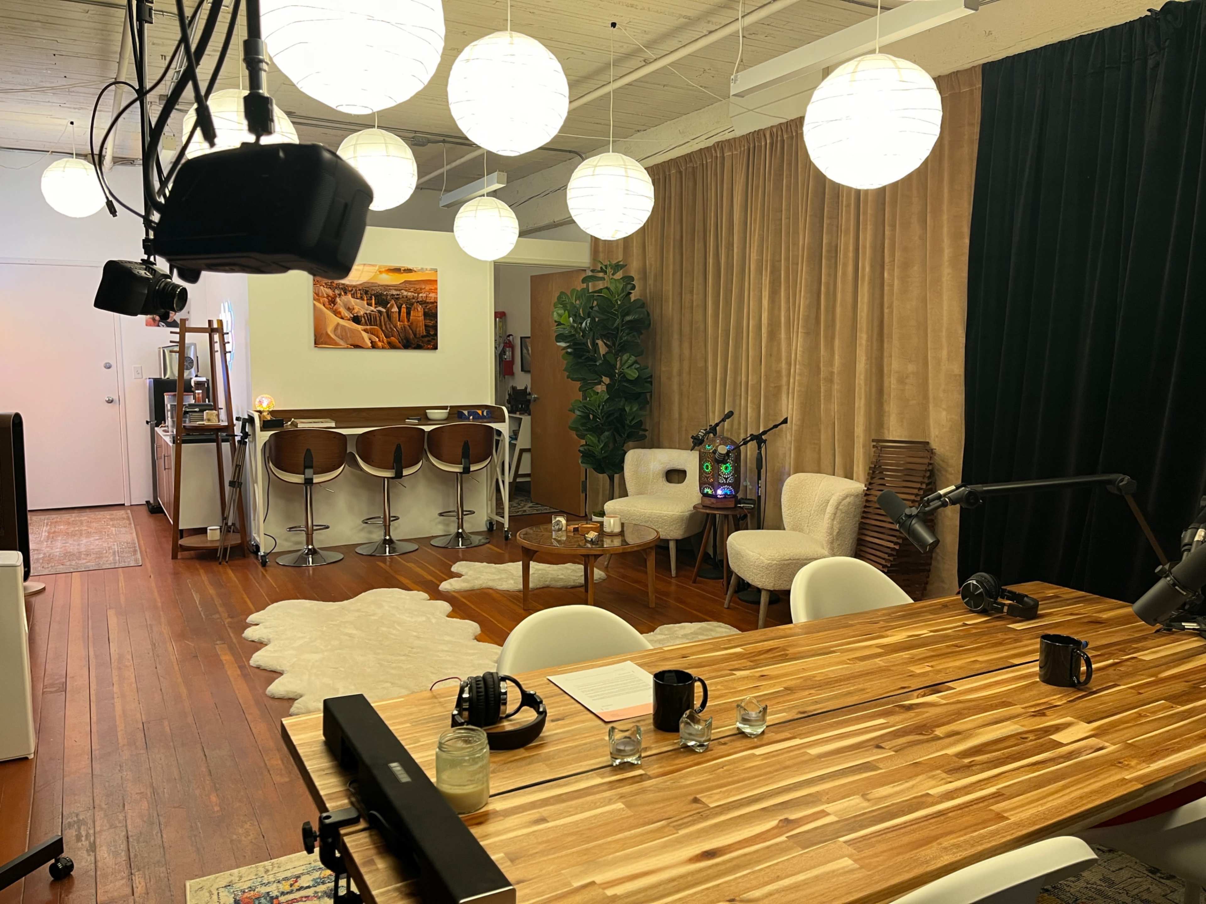 The image shows a modern recording studio featuring a wooden table, microphones, and cozy seating arranged in a well-lit room with pendant lights and a bar area in the background.
