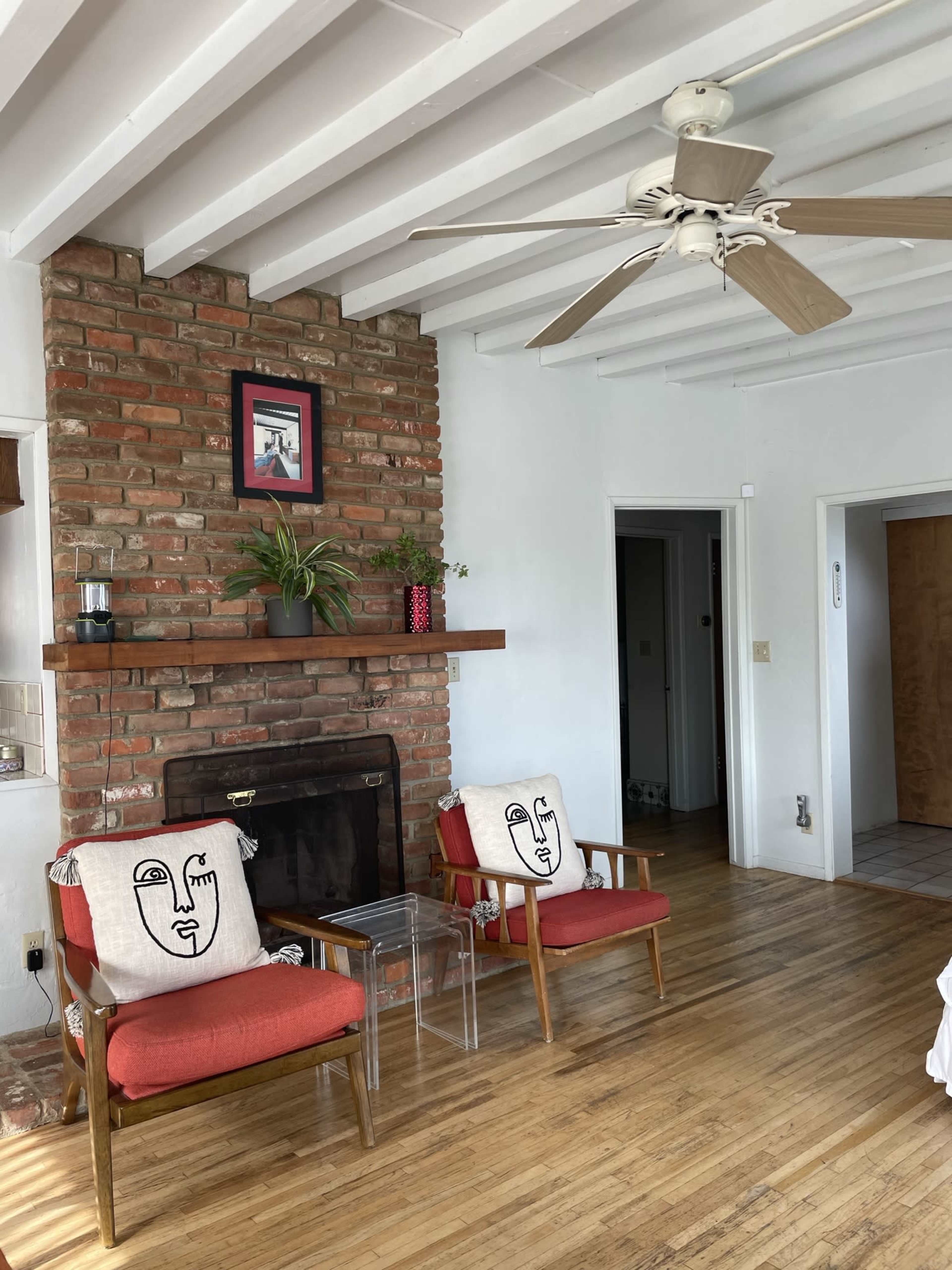 A living room with two red chairs, a brick fireplace, and a ceiling fan, complemented by a potted plant and wall art.