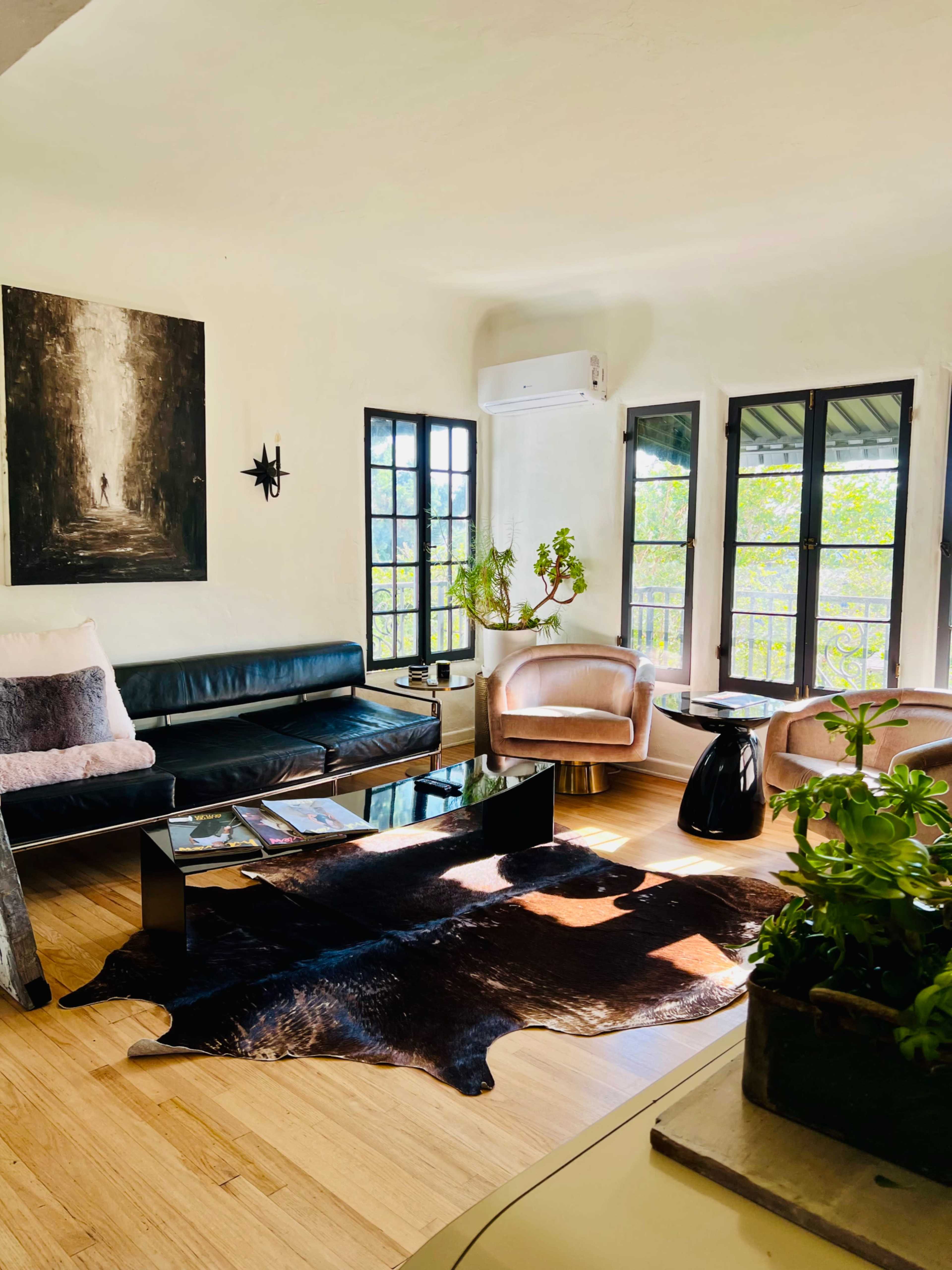 The image shows a modern living room with a black leather sofa, two pink armchairs, a glass coffee table on a cowhide rug, and large windows allowing natural light to fill the space.