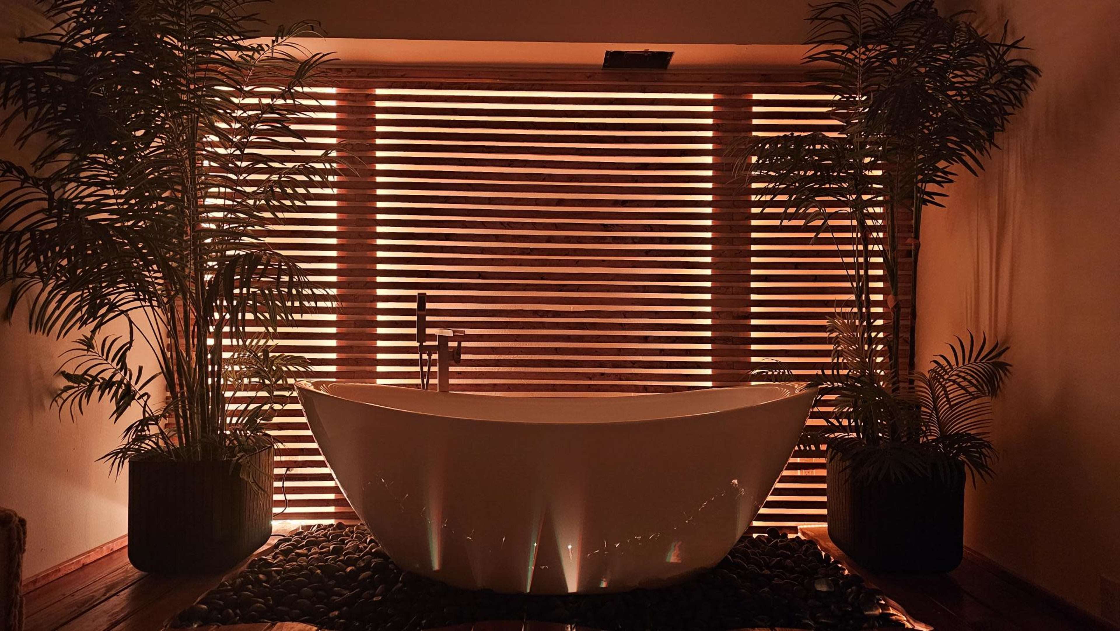 A white freestanding bathtub is positioned in front of a slatted wooden screen illuminated by warm light, surrounded by potted plants and dark stones on the floor.