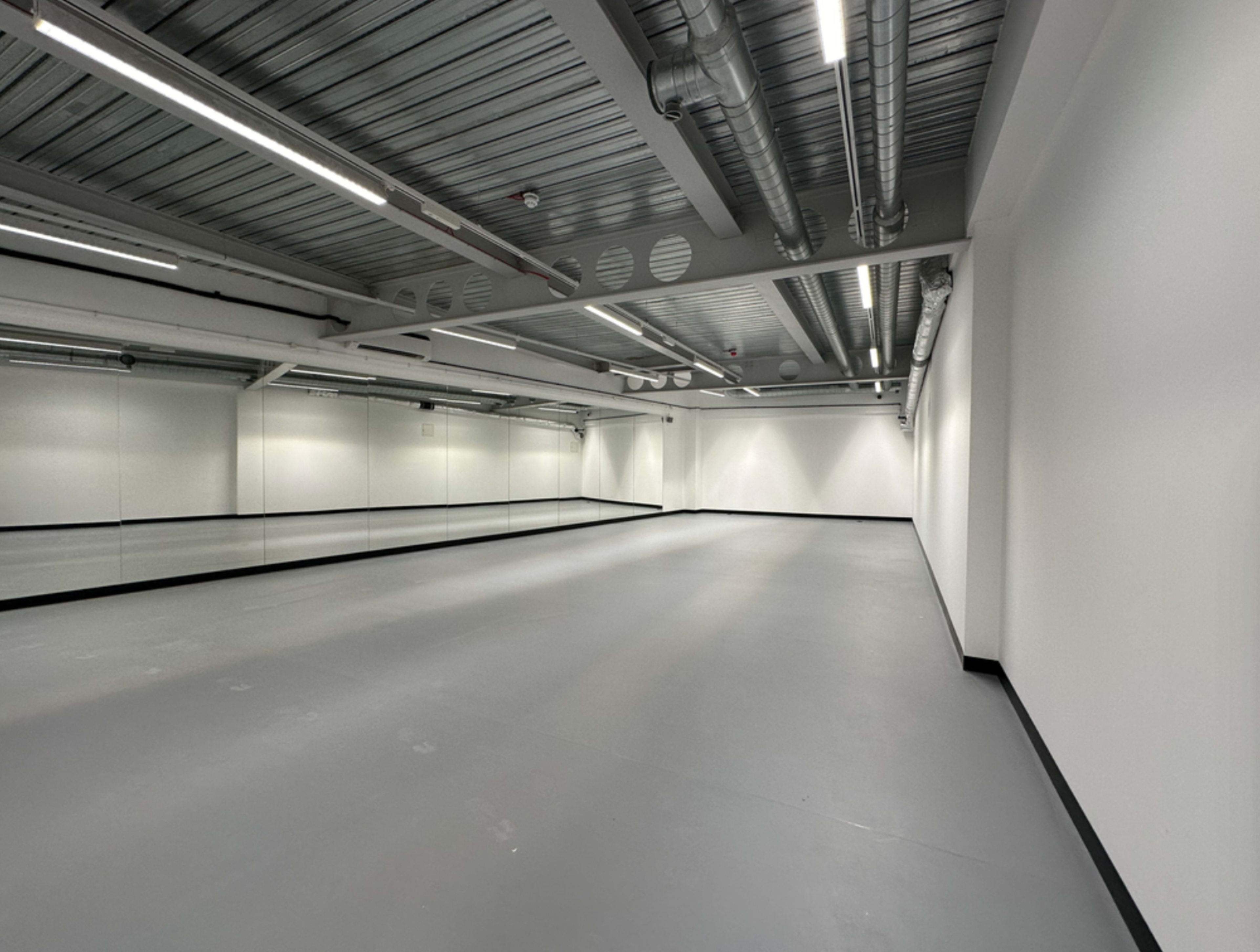 A spacious, empty room with gray flooring and a wall of mirrors under exposed ceiling infrastructure.