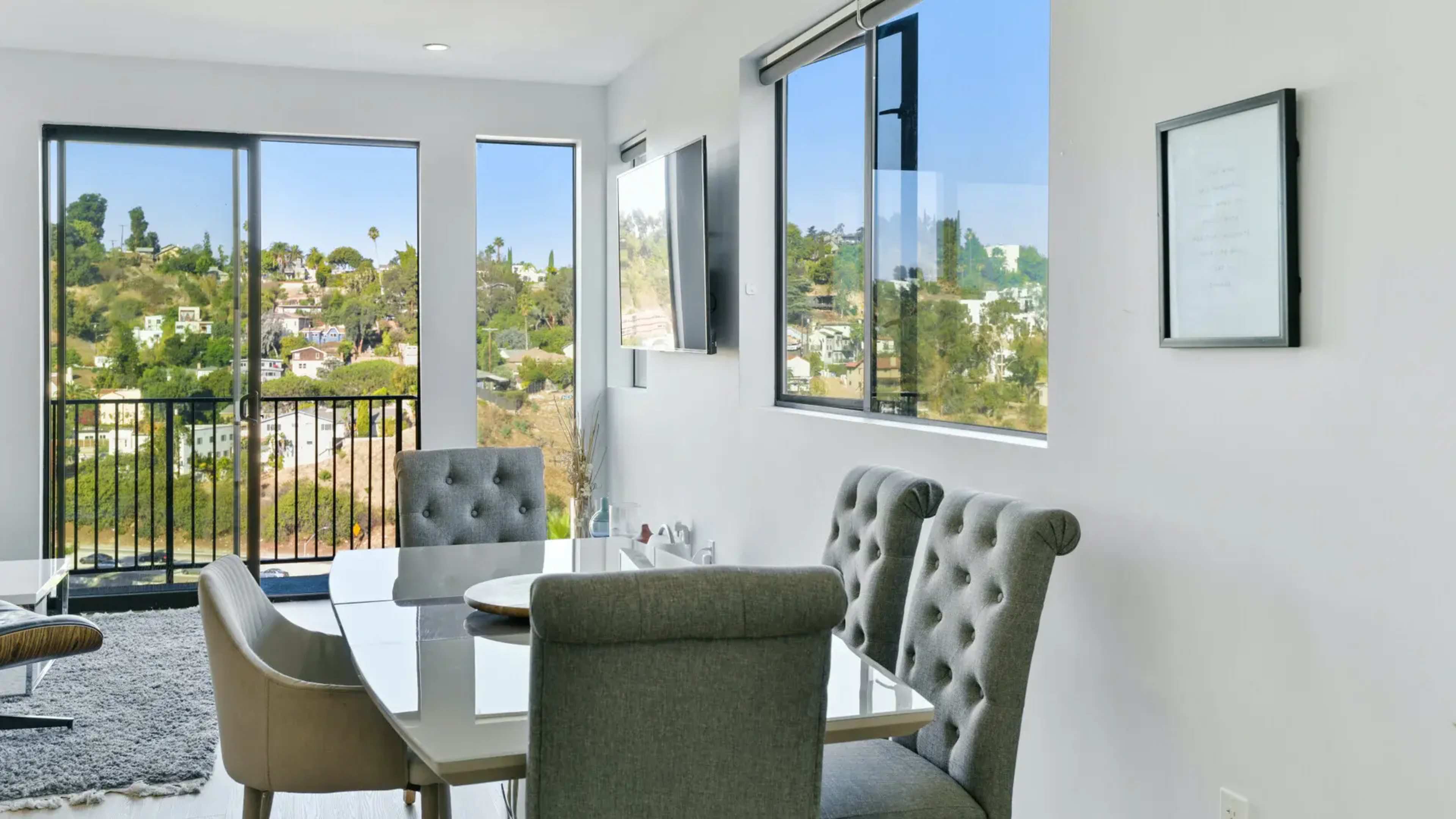 The image shows a modern dining area with a glass table and upholstered chairs, overlooking a hillside view through large windows.