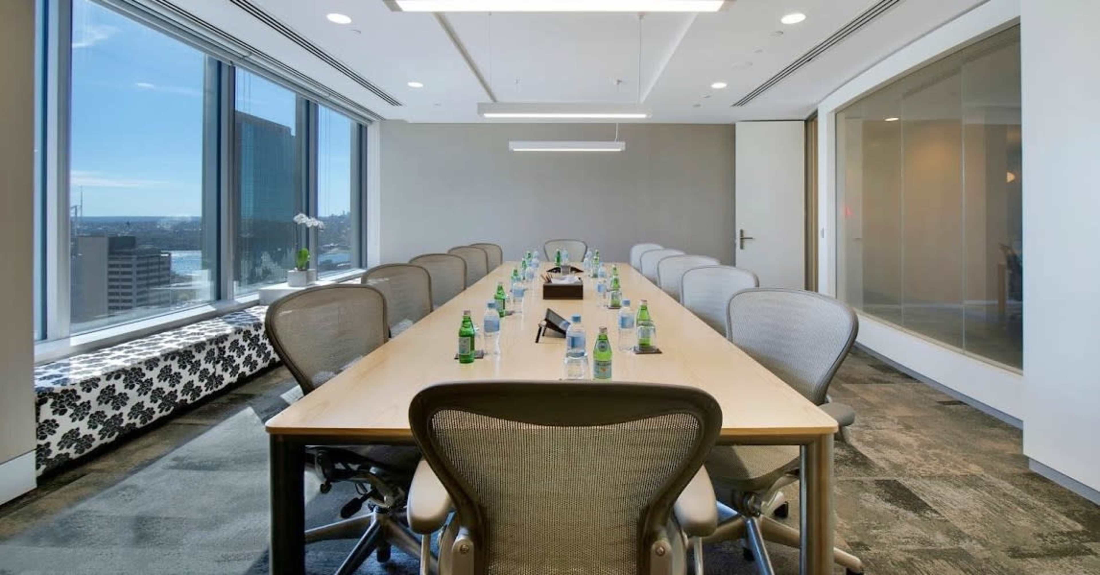 A long conference table with chairs is arranged in a modern office meeting room, featuring large windows that provide a view of the cityscape.