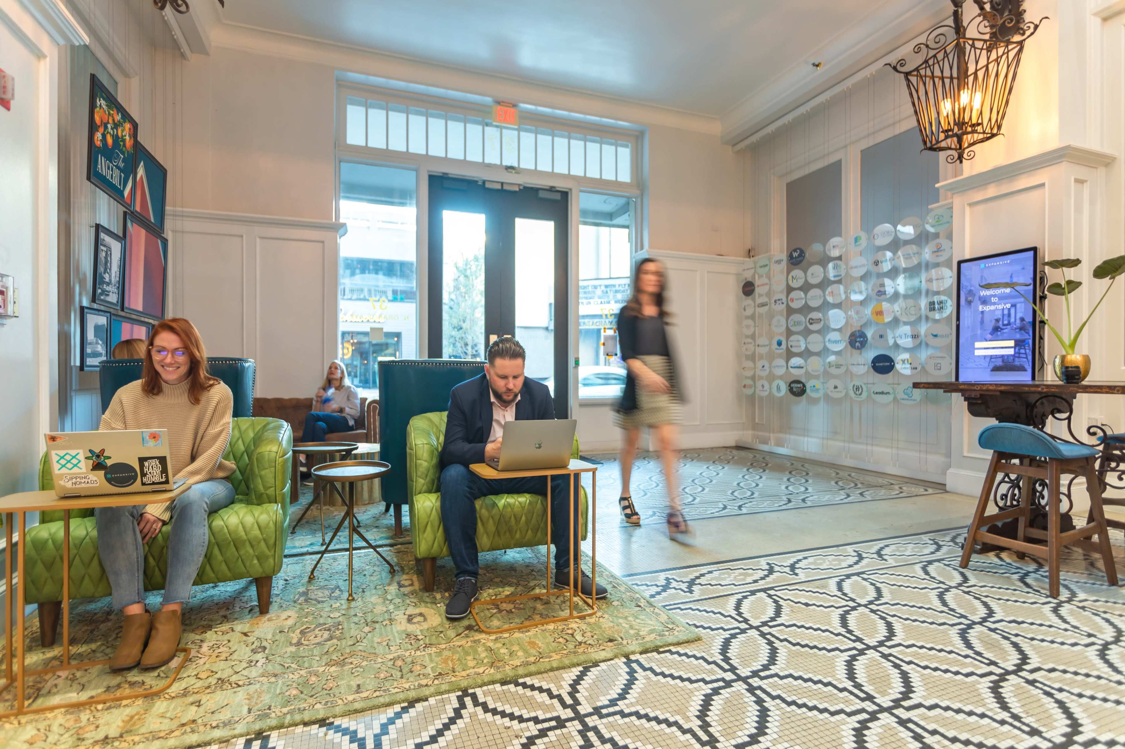 Two individuals are seated in green chairs working on laptops in a brightly lit lobby with a patterned floor and decorative elements.