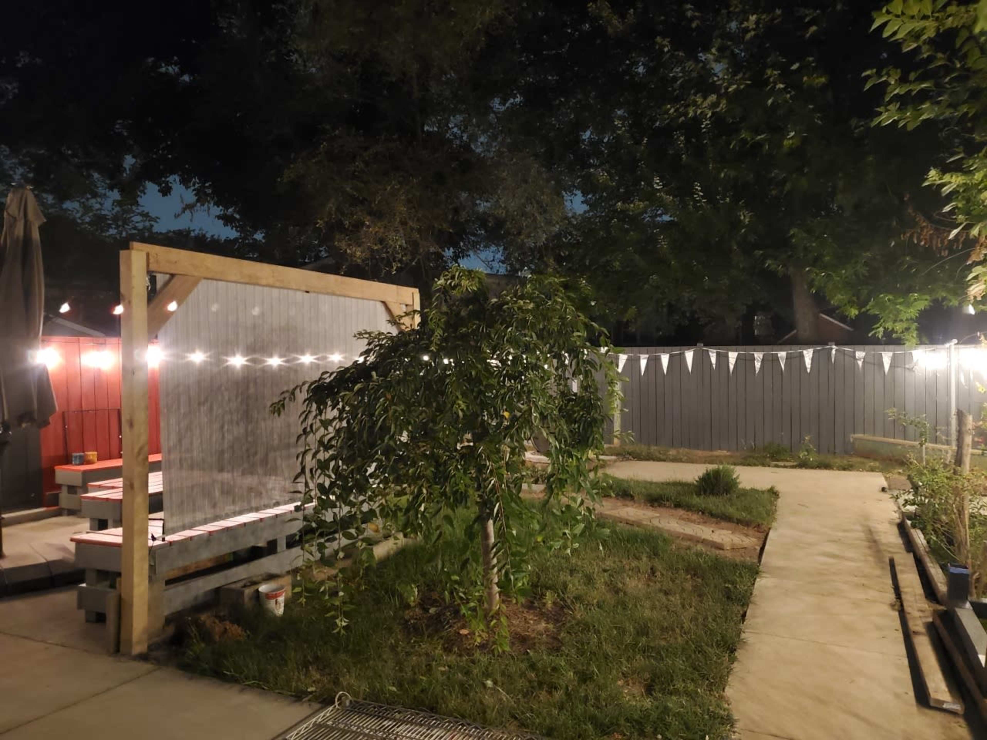 The image shows a backyard with a wooden structure adorned with string lights, a small tree, and a paved walkway bordered by grass and plants.