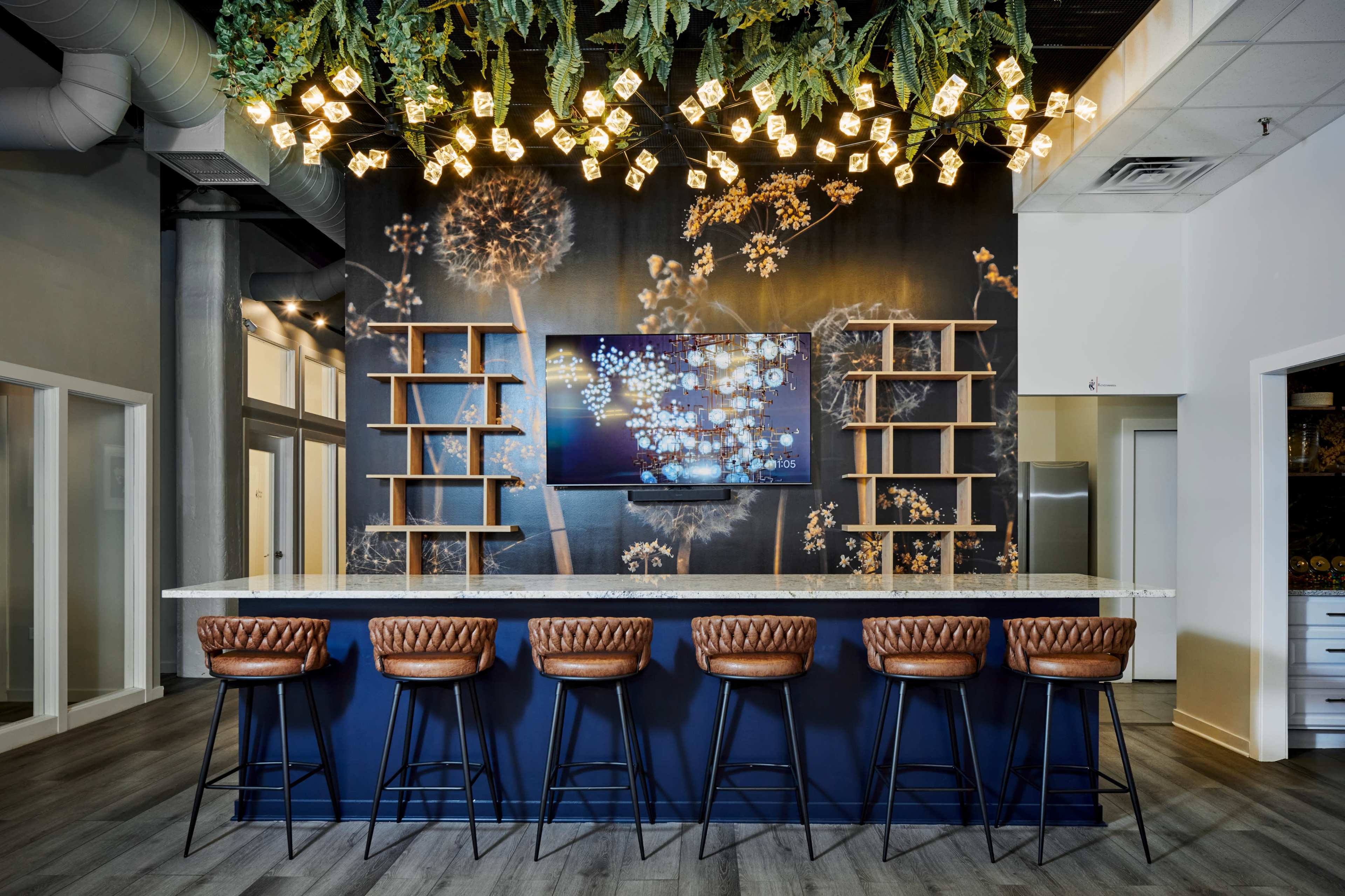 A modern bar area with a granite countertop, leather bar stools, and a decorative wall featuring a television and shelves adorned with plants.