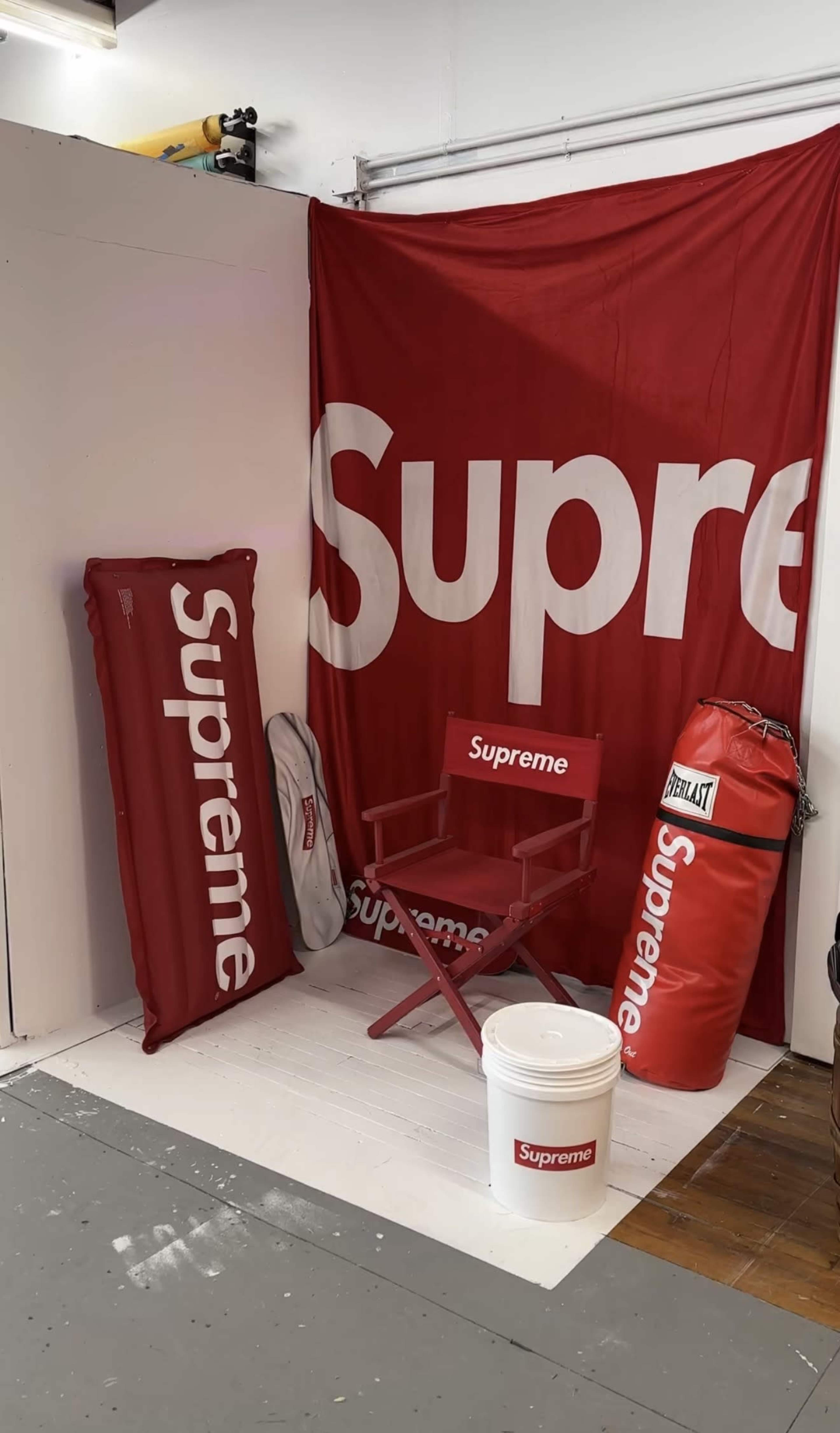 The image shows a corner display featuring various red and white Supreme-branded items, including a chair, a storage bucket, and a punching bag against a large Supreme banner.