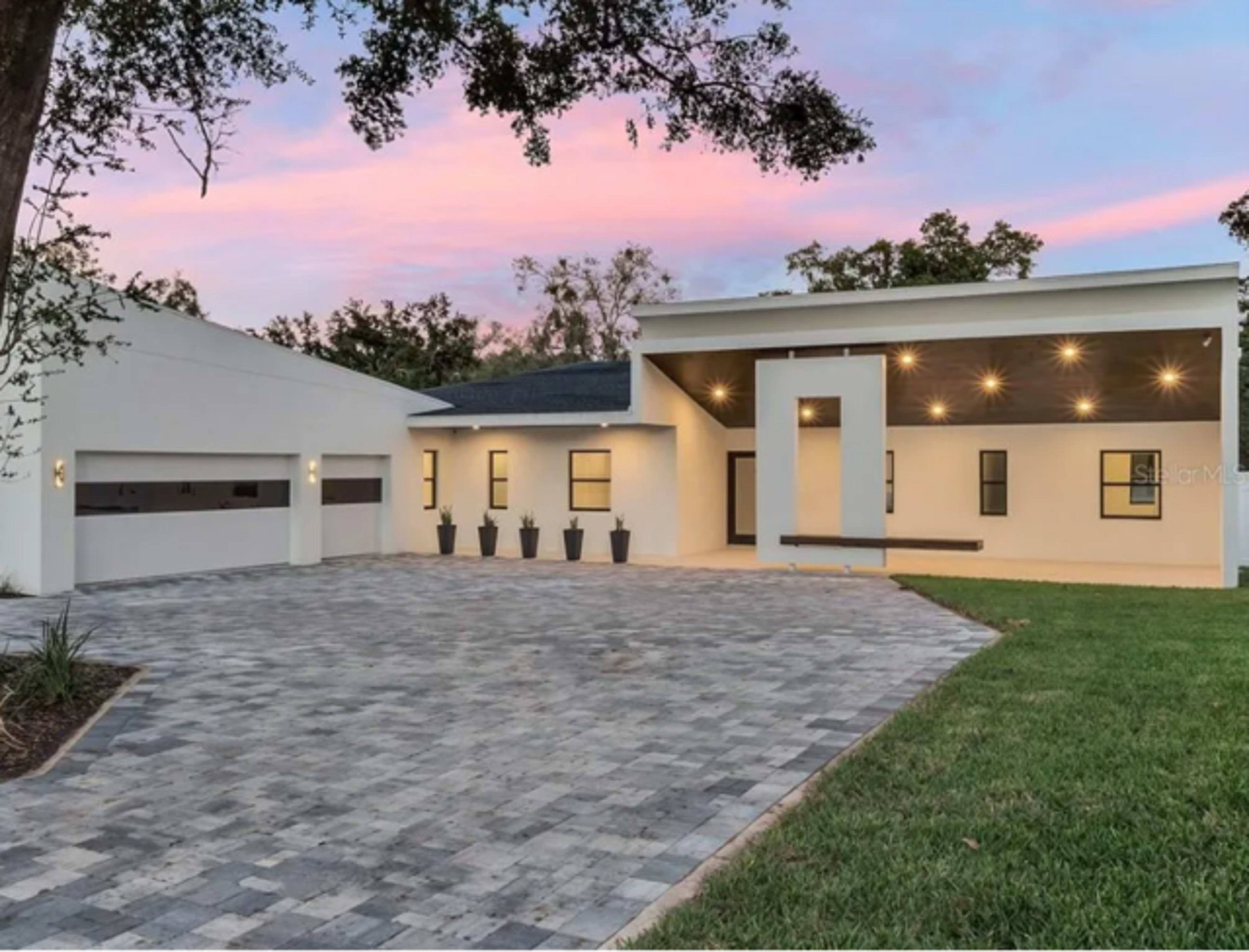 The image shows a modern house with a large driveway, featuring a landscaped front yard and several potted plants near the entrance.